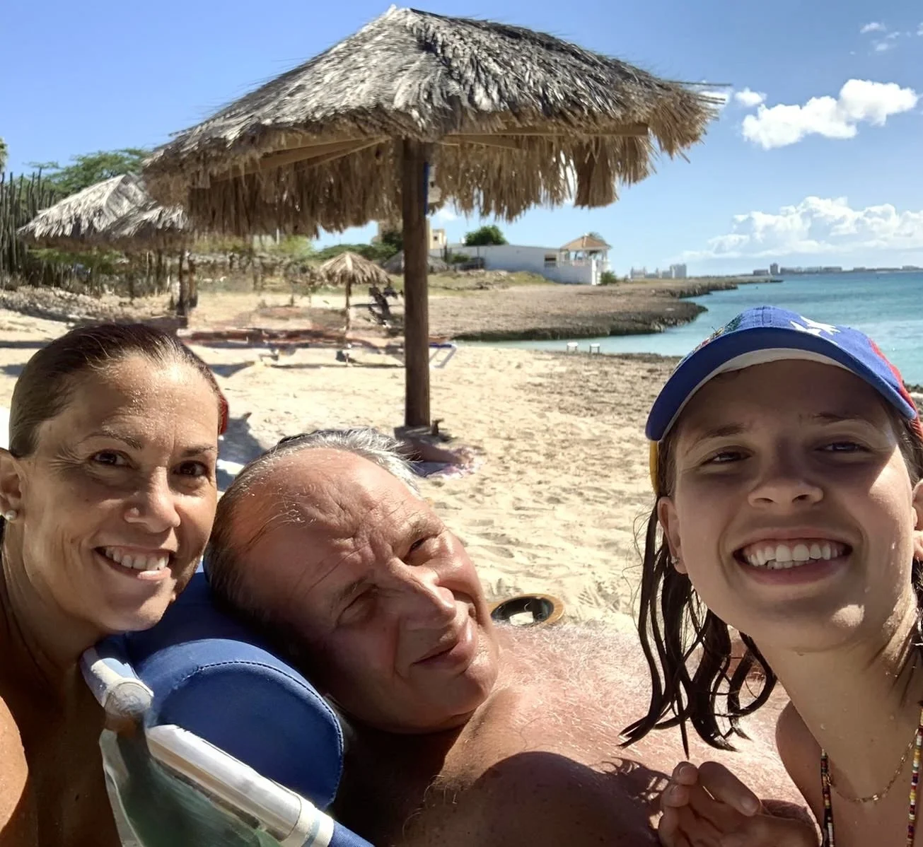A family of three taking a selfie on a sunny beach with straw umbrellas, sandy shore, and ocean in the background.