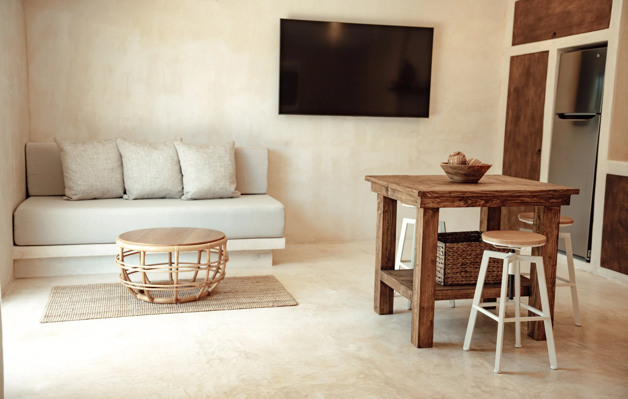 Minimalist living room with beige walls and floor, gray sofa with three cushions, round rattan coffee table, wooden dining table with a bowl of decorative balls, white stools, and a large flat-screen TV mounted on the wall.