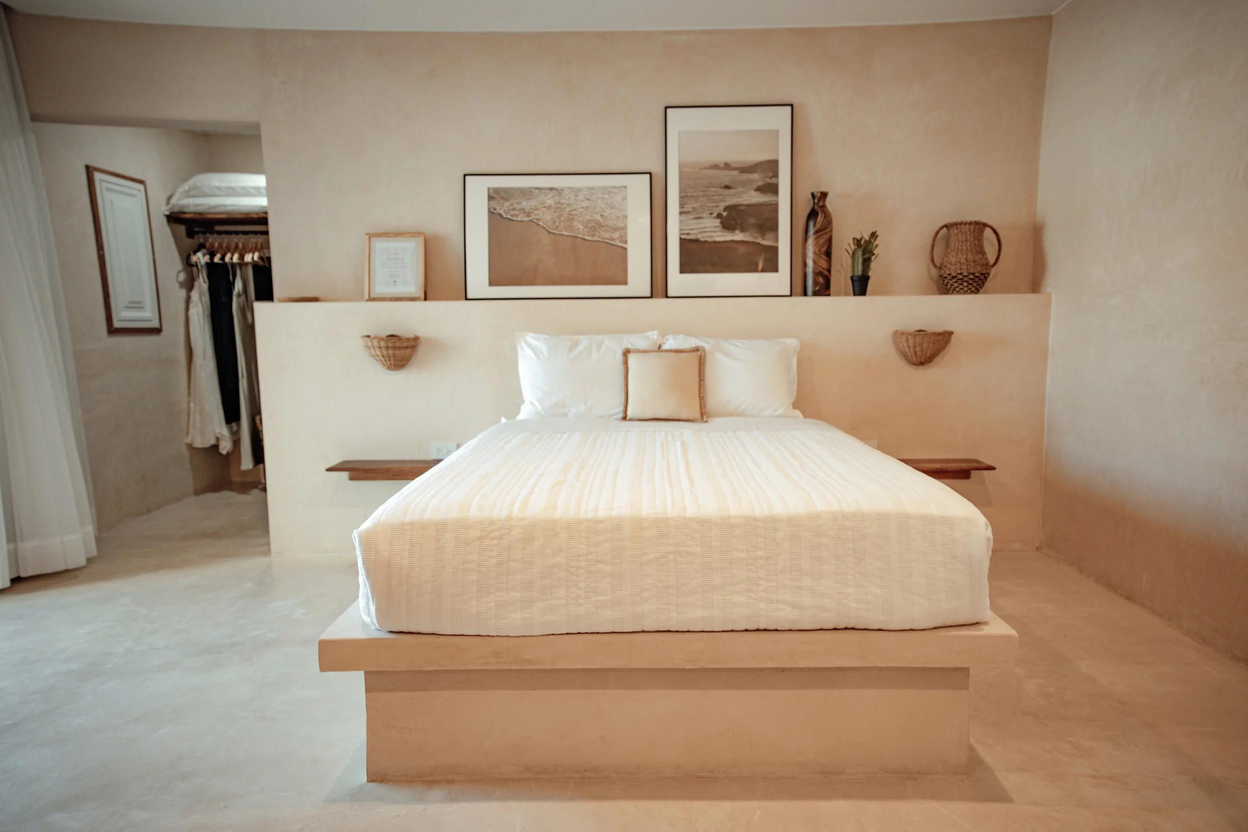 A cozy bedroom with a minimalist design, featuring a large bed with white bedding and pillows, decorated with framed artwork of beach landscapes on the beige wall behind, and accented with natural wicker and wooden decor elements.