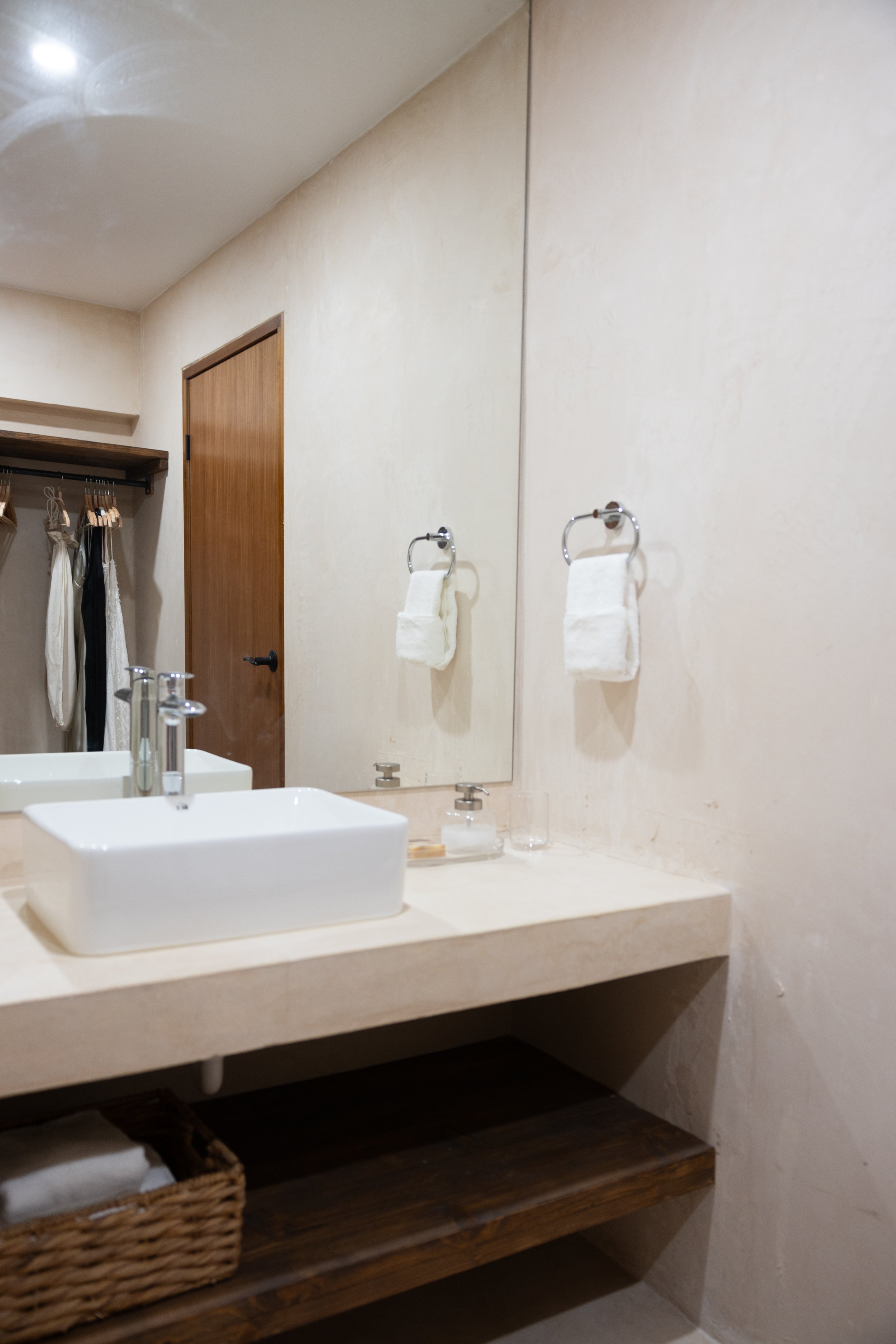 A bathroom sink with a white rectangular vessel, a glass soap dispenser, and a clear glass bottle in front of a large mirror. Towel rings with white towels hang on the wall, and a wooden door and closet with hanging clothes are in the background.