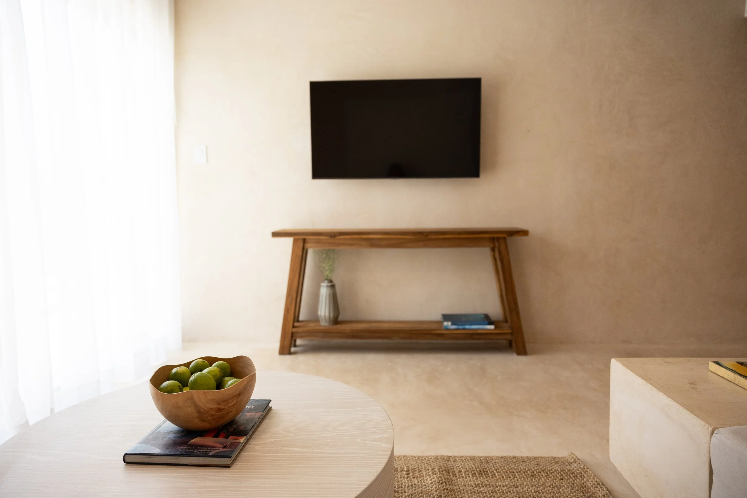 Living room with a wall-mounted TV, a wooden console table with a vase and books, a round table with a bowl of green limes, a magazine, and a beige armrest in the foreground.