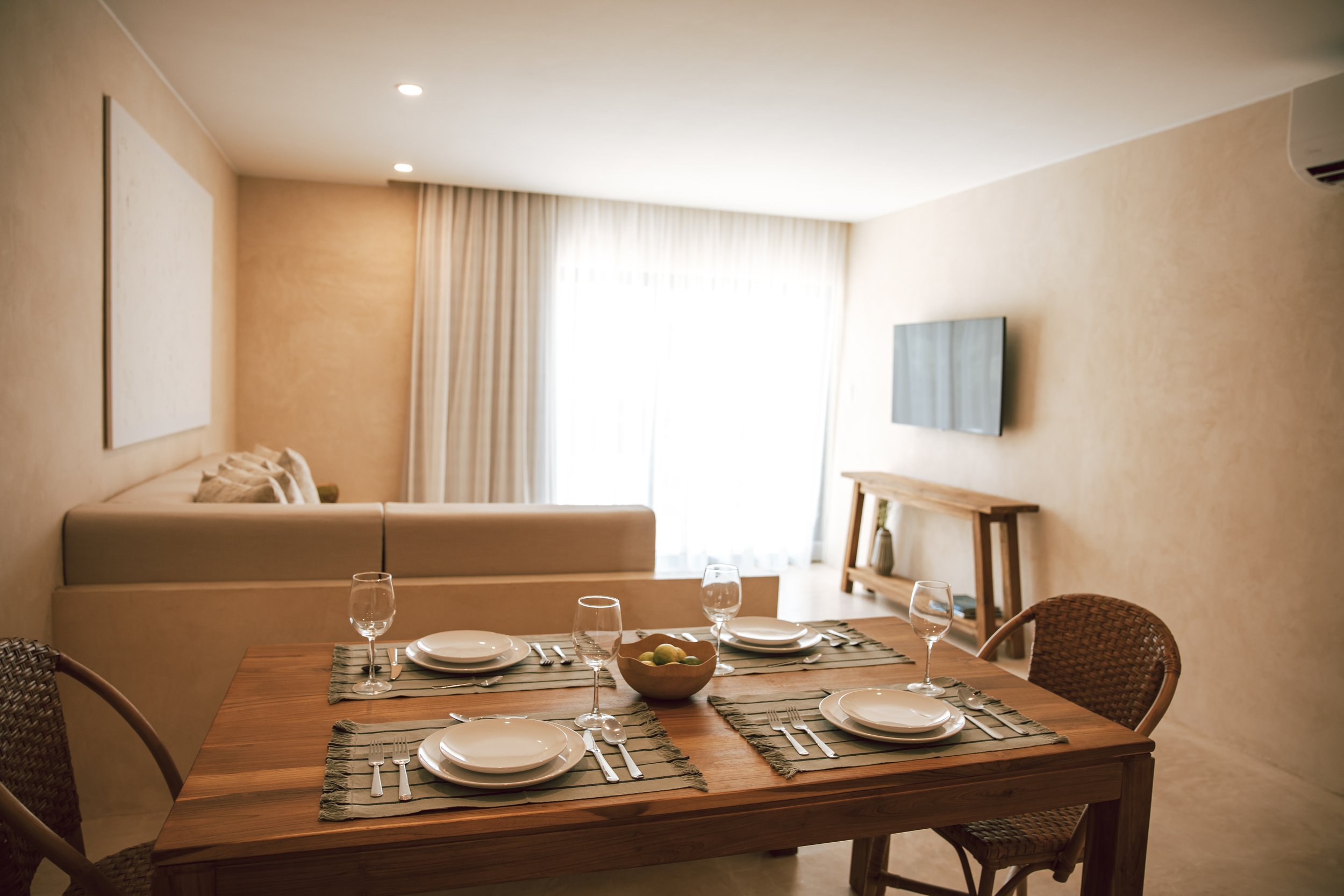 Dining area with a wooden table set with four place settings, wine glasses, a bowl of lemons, wicker chairs, and a living room with a beige sofa, curtains, and a wall-mounted TV in the background.