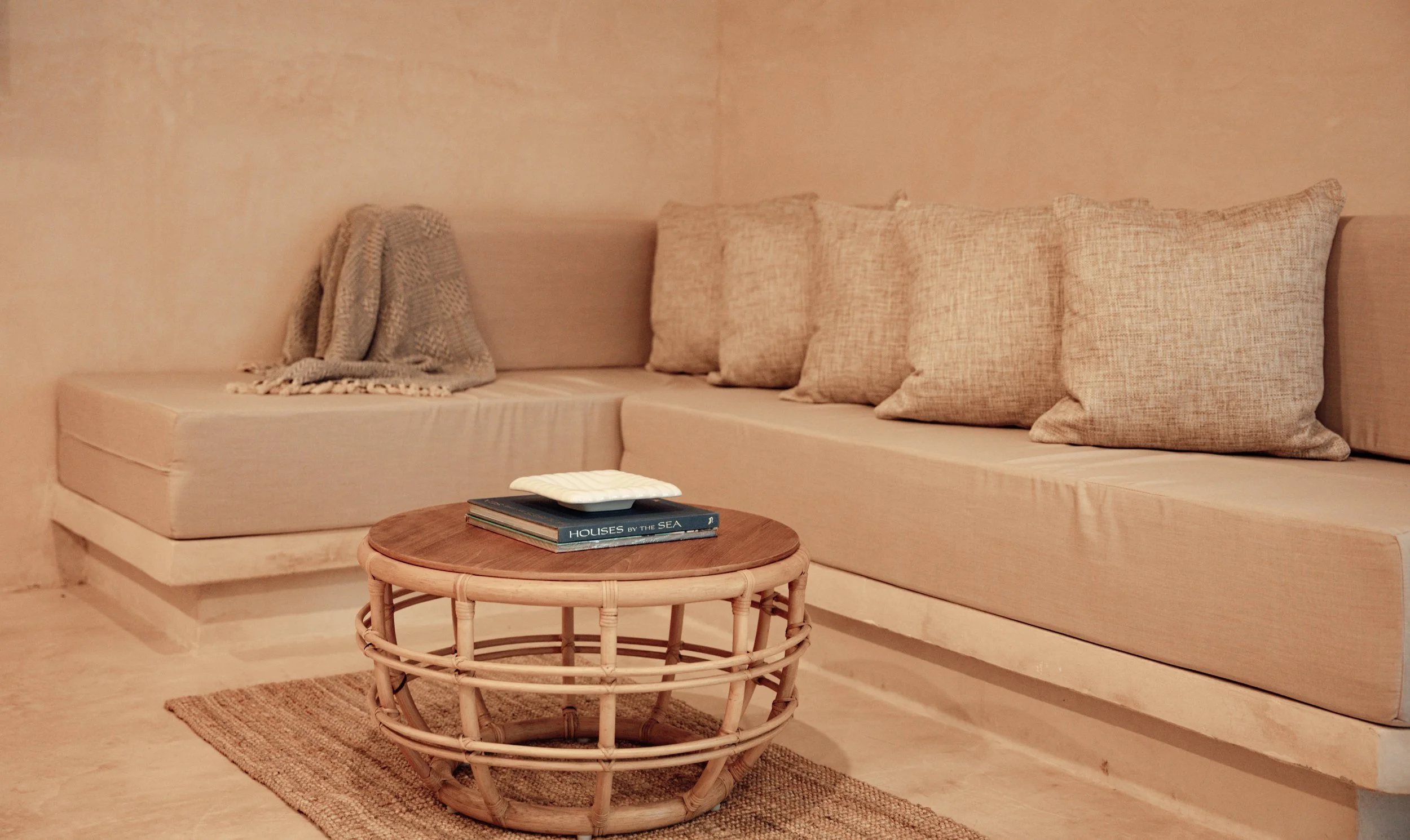 Cozy beige living room corner with a sectional sofa, beige cushions, a gray throw blanket, a round rattan coffee table with books and a decorative item, and a woven rug.