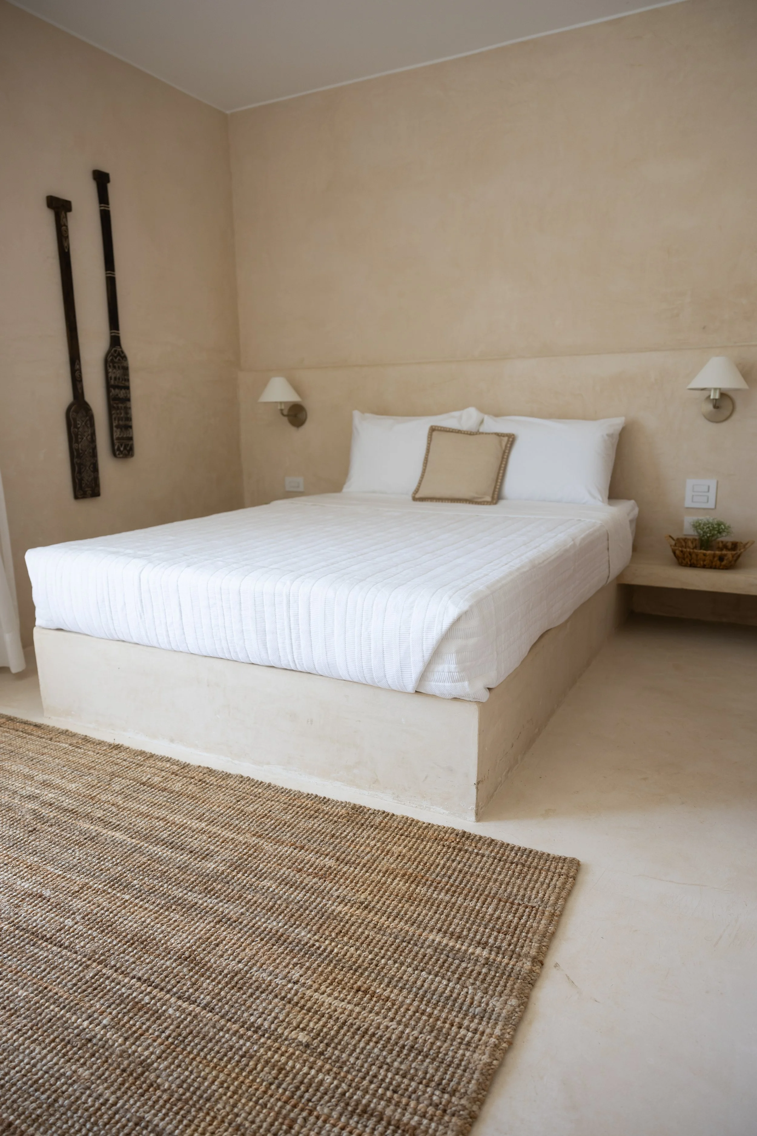 A neatly made bed in a minimalist bedroom with beige walls, wall-mounted lamps, and decorative paddles hanging on the wall.
