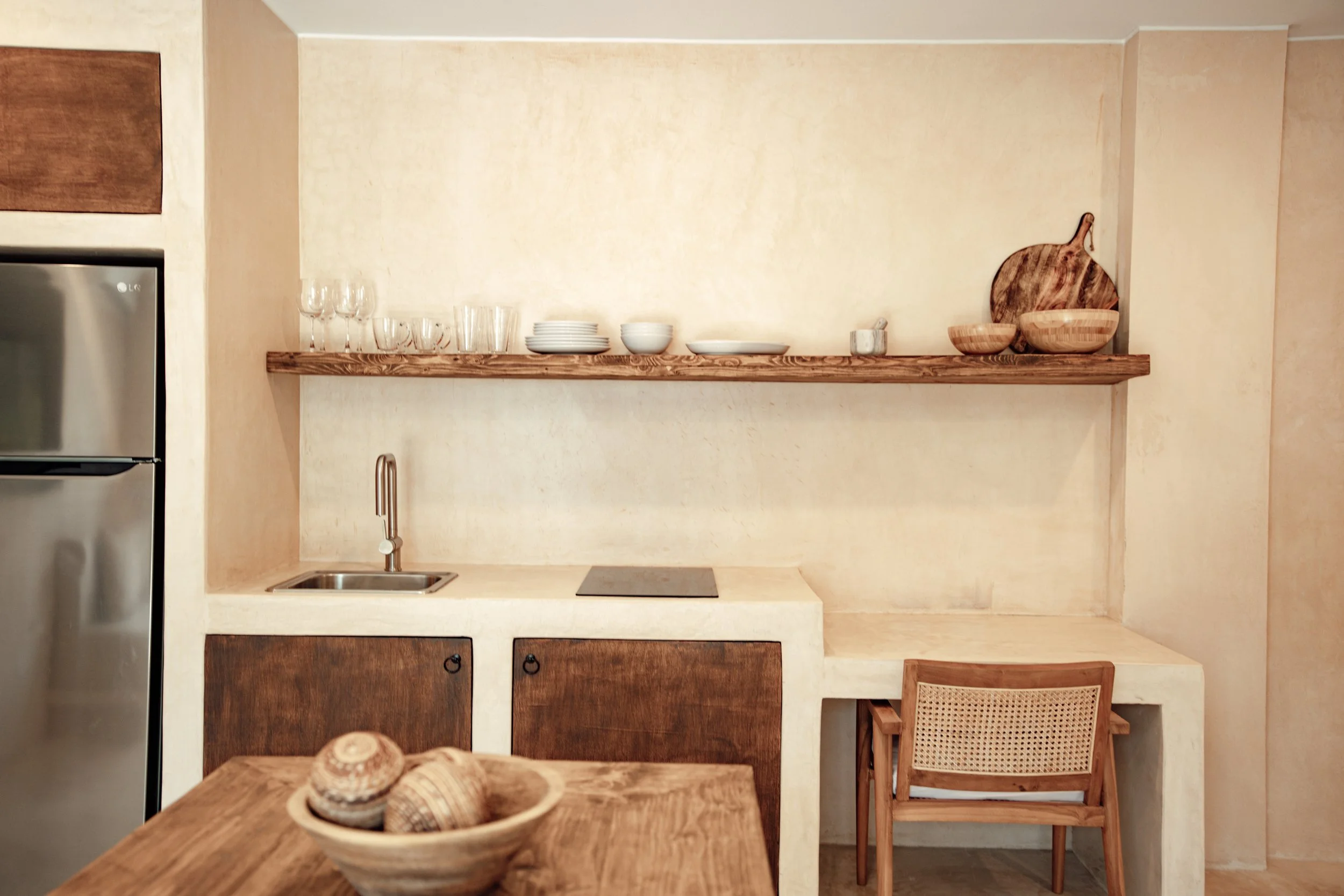 A minimalist kitchen with a wooden countertop, open shelf with glassware and bowls, small sink, and a wooden chair and table with seashells in a bowl.