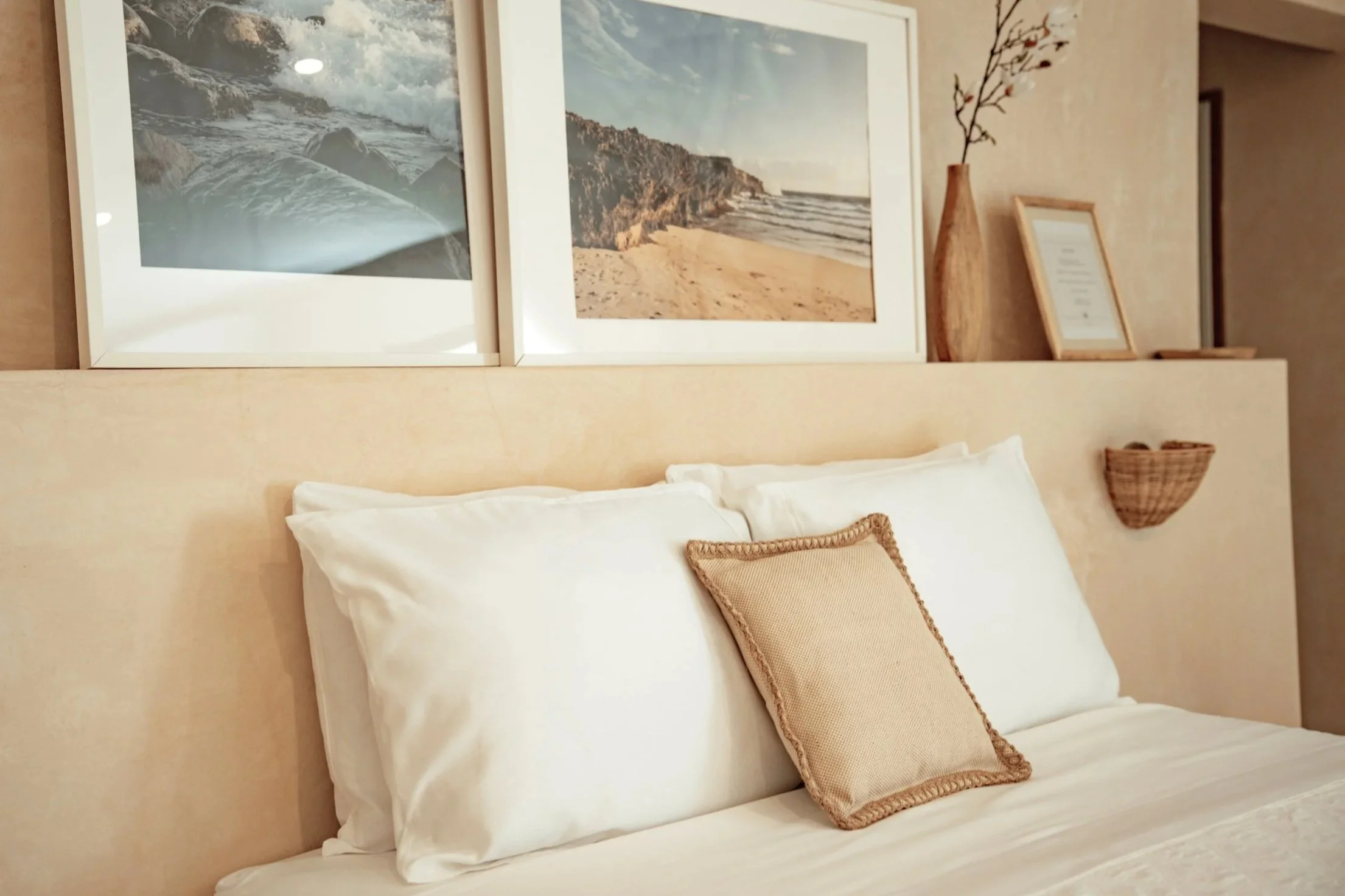 A cozy bedroom with a bed featuring white pillows and a beige decorative pillow. Above the bed is a beige headboard with artwork and decorative items, including framed beach photos, a framed document, a tall brown vase with branches, and a small wicker wall shelf.