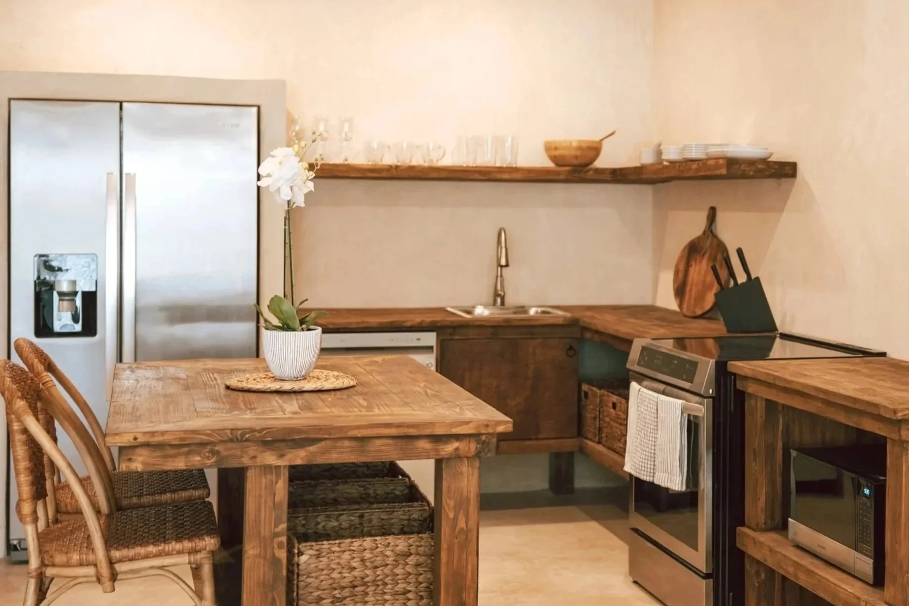 A rustic kitchen with wooden countertops, a stainless steel refrigerator, a stainless steel oven, and open shelves. There is a small dining table with chairs, a potted orchid on the table, and various kitchen utensils and dishes displayed around.