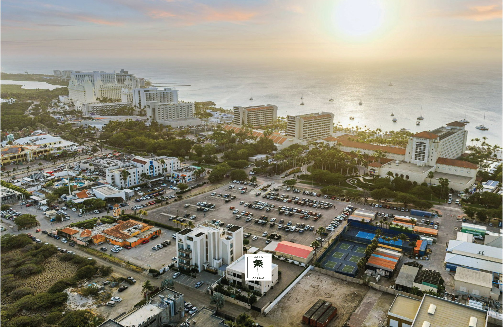 Aerial view of a coastal cityscape at sunset, featuring a parking lot, tennis courts, various buildings, trees, and the ocean with sailboats in the background.