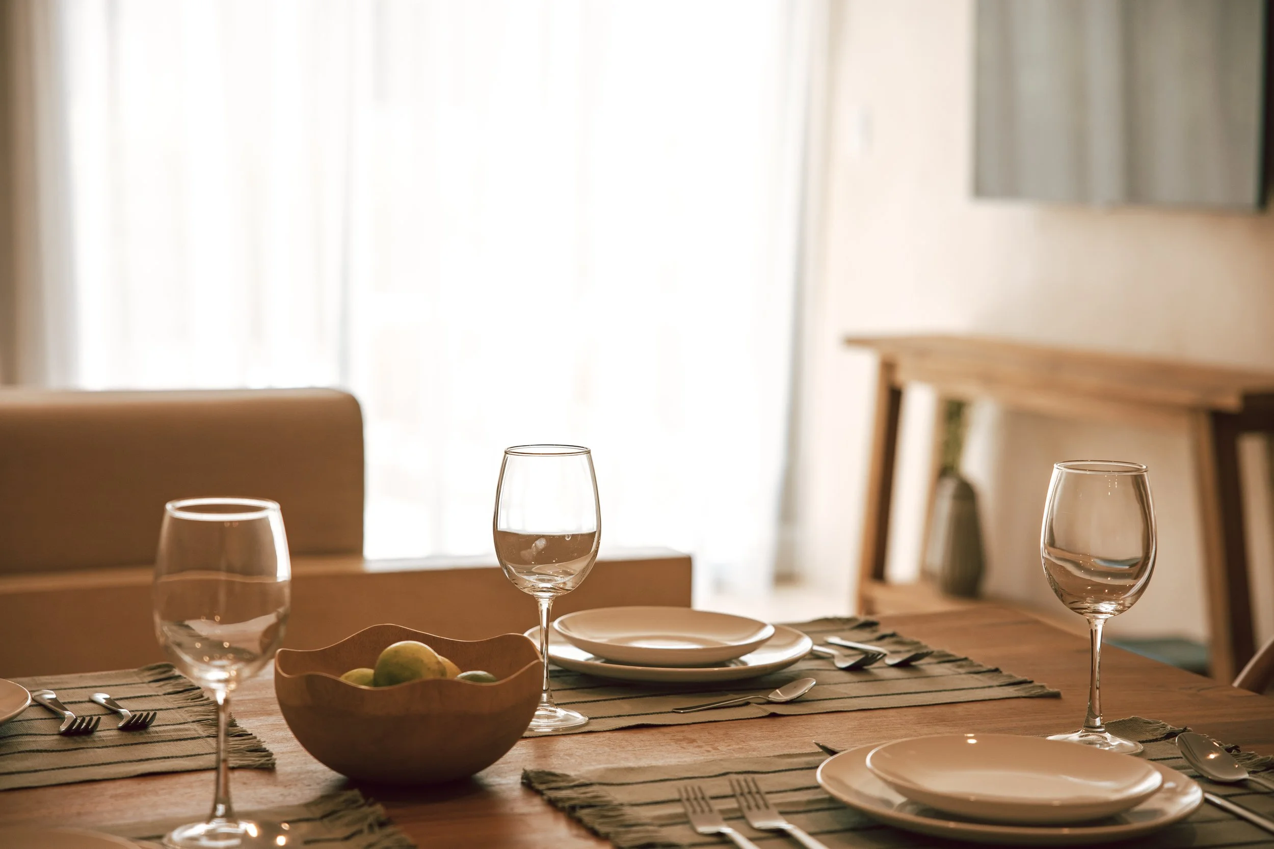 A dining table set with four plates, four forks, and four glasses of water, with a wooden bowl of green apples in the center, in a room with sheer curtains letting in natural light.