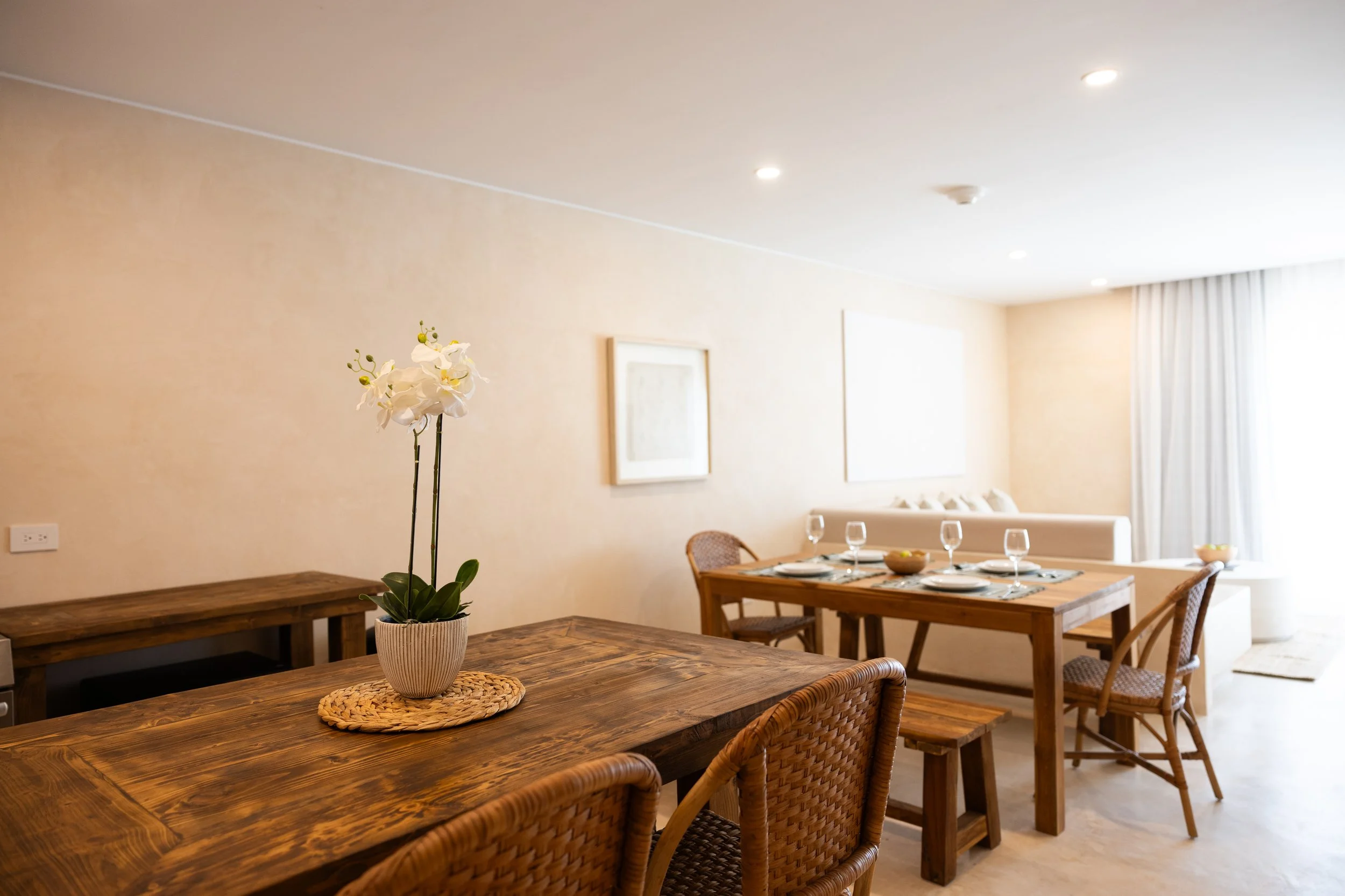 Dining room with a wooden table set with plates and glasses, and an orchid in a pot as a centerpiece, in front of a beige wall with framed artwork and a window with curtains