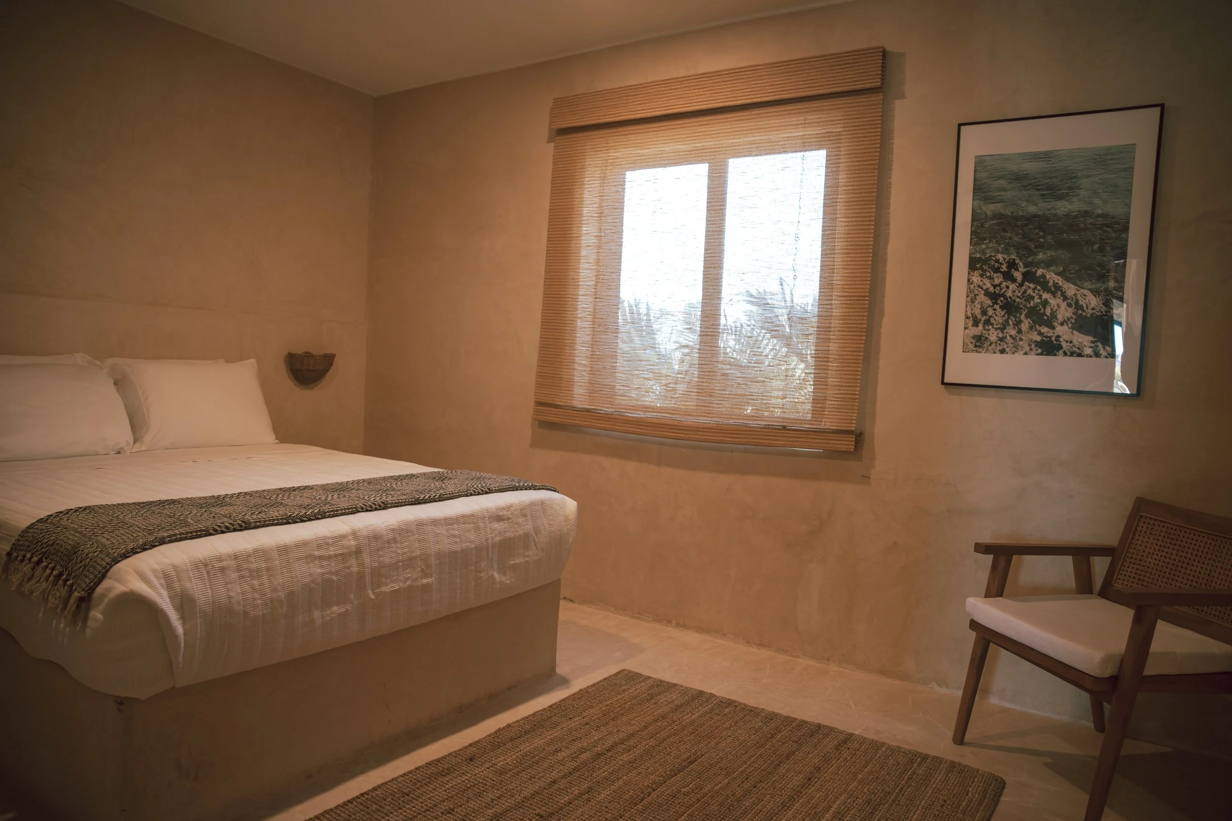 A bedroom with beige walls, a king-size bed with white linens and a brown textured blanket, a window with bamboo blinds, a framed landscape photograph on the wall, a wooden chair with a white cushion, and a neutral-colored rug.