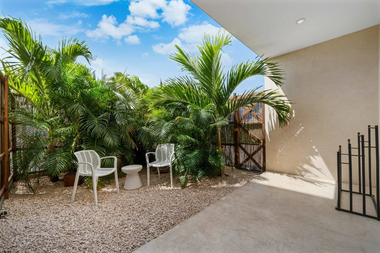 A small outdoor patio with white chairs, a small white side table, and lush green tropical plants against a blue sky with light clouds.