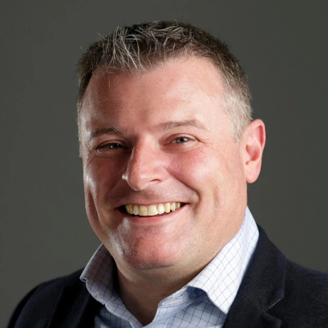 Headshot of smiling middle-aged man with short, grayish hair, wearing a dark blazer and white checkered shirt.
