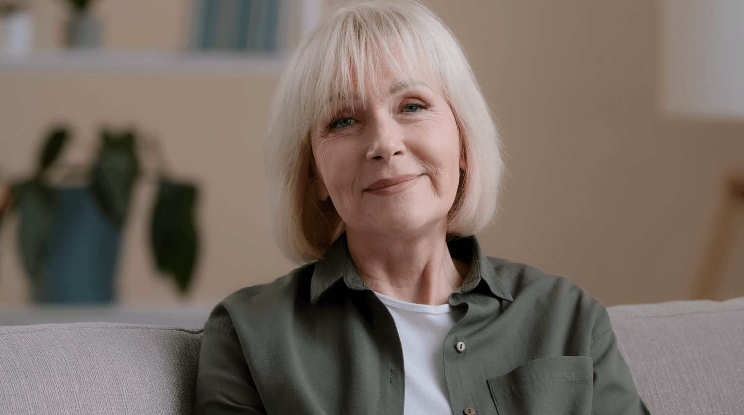 A smiling middle-aged woman with blonde hair sitting on a beige couch in a cozy living room.