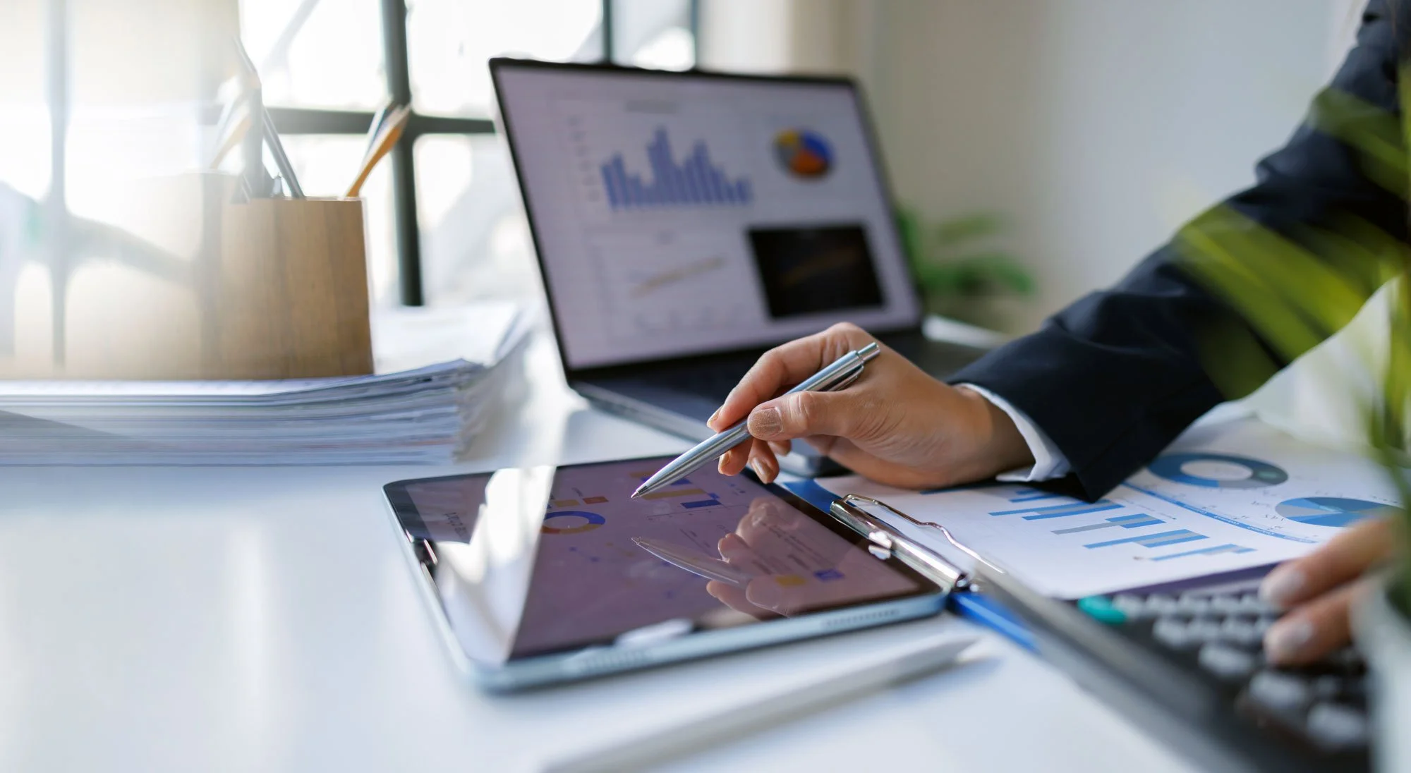 Person in business attire using a stylus on a tablet with financial charts, with a laptop displaying graphs and pie charts, on an office desk with documents and a pen holder.