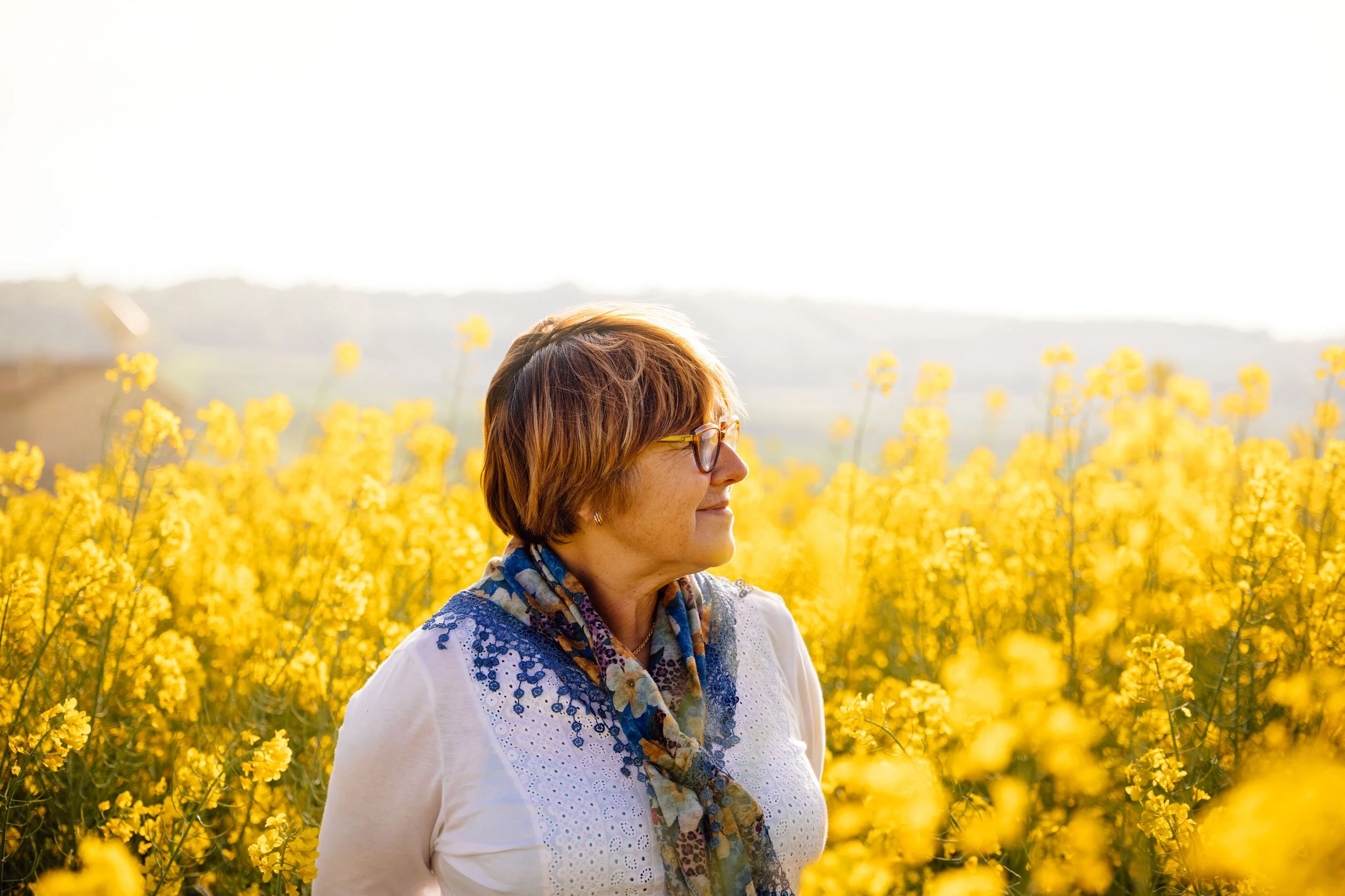 A woman with short hair and glasses standing in a bright yellow flower field during golden hour sunlight.