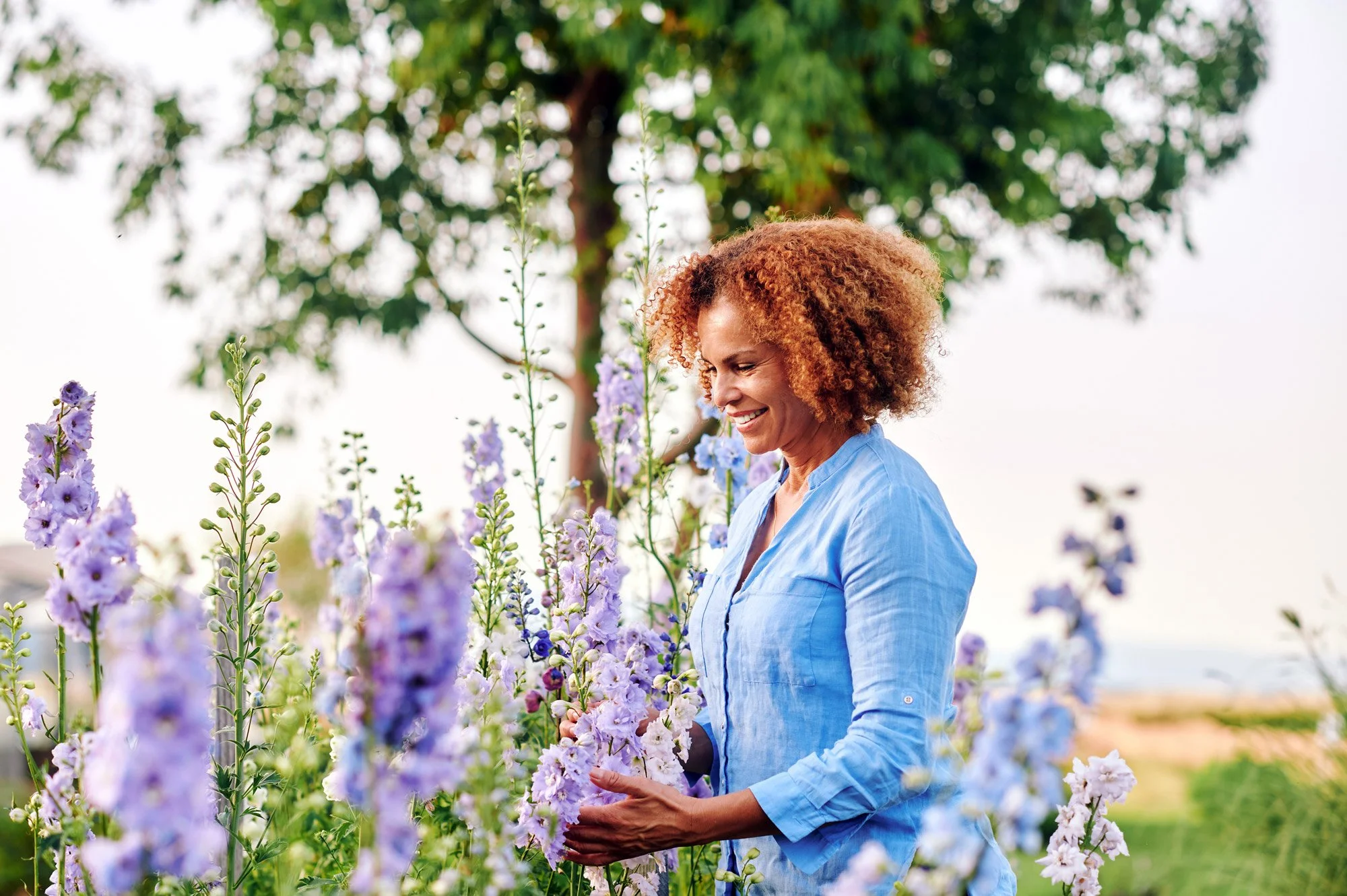 A woman with curly red hair wearing a light blue shirt is smiling while standing among purple and white flowers outdoors.