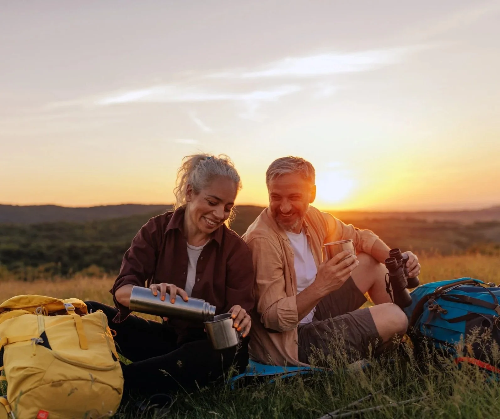 A happy middle-aged couple sitting on the grass in a field during sunset, preparing hot drinks with thermoses and mugs, surrounded by backpacks, enjoying outdoor camping.