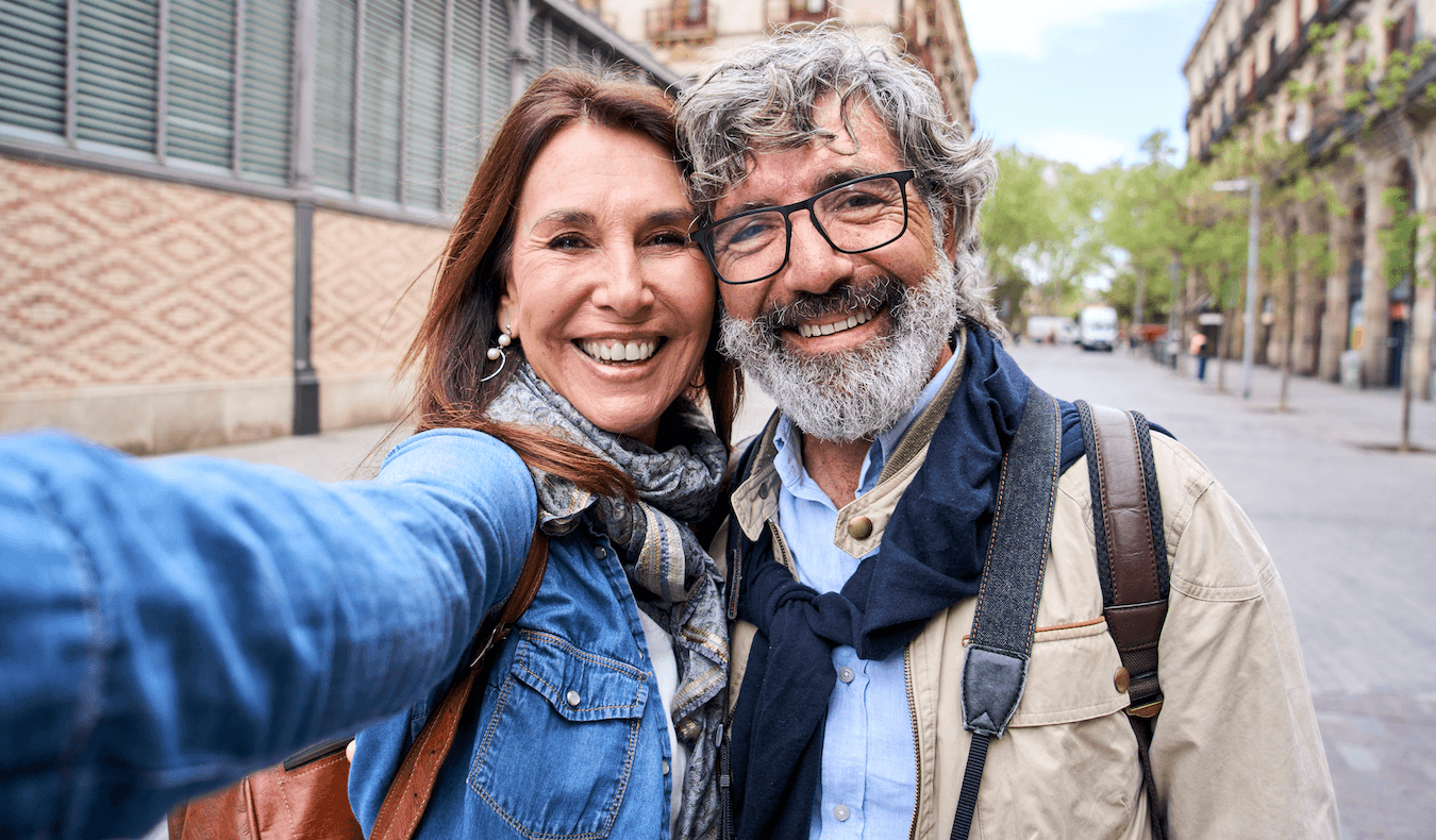 A smiling couple taking a selfie on a city street, with buildings and trees in the background.