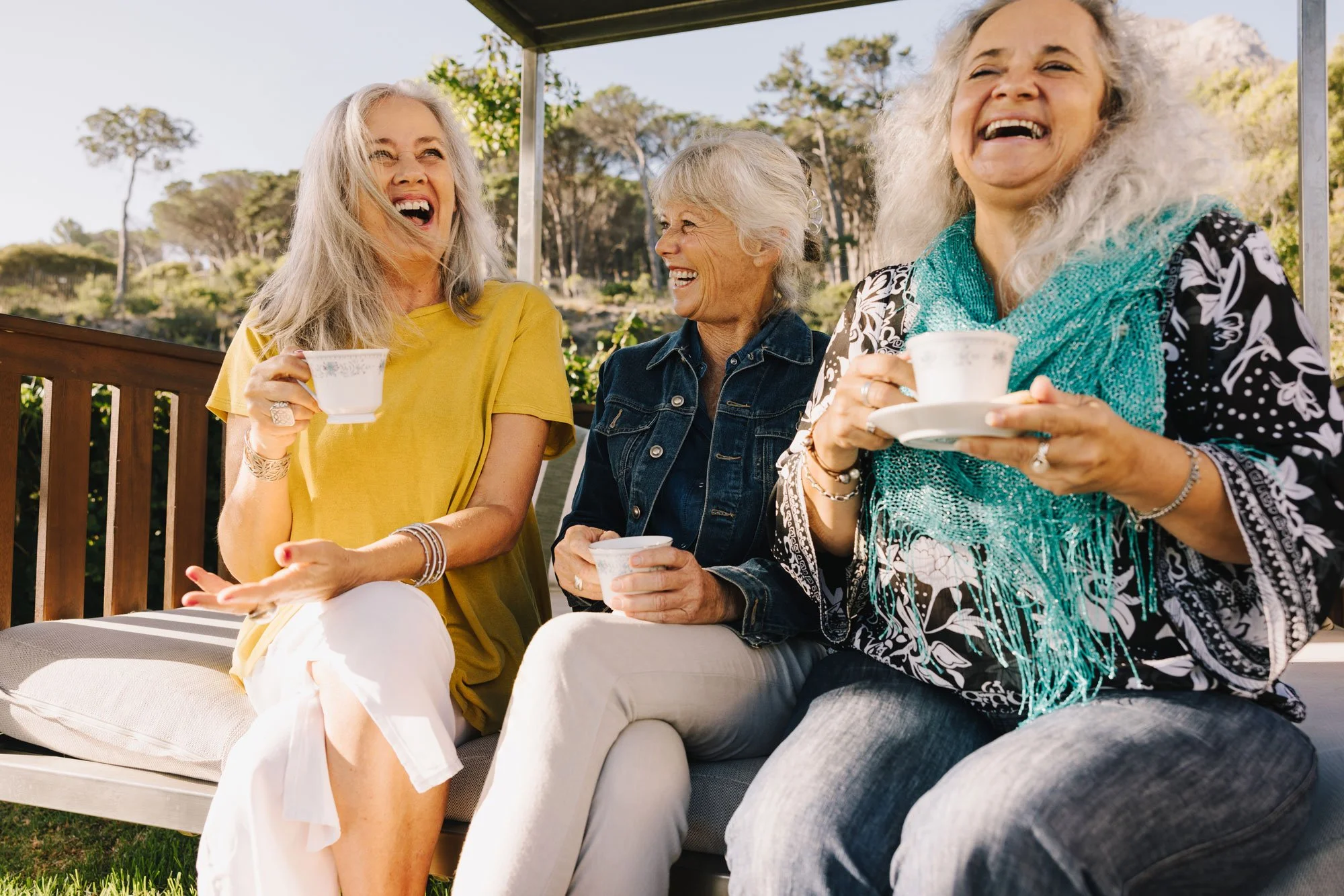 Three older women sitting on a bench outdoors, laughing and enjoying tea or coffee, with trees and a rocky hill in the background.