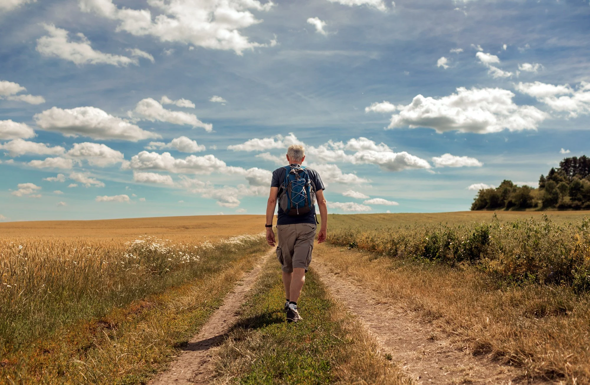 Man walking on a dirt trail through open fields under a partly cloudy sky.