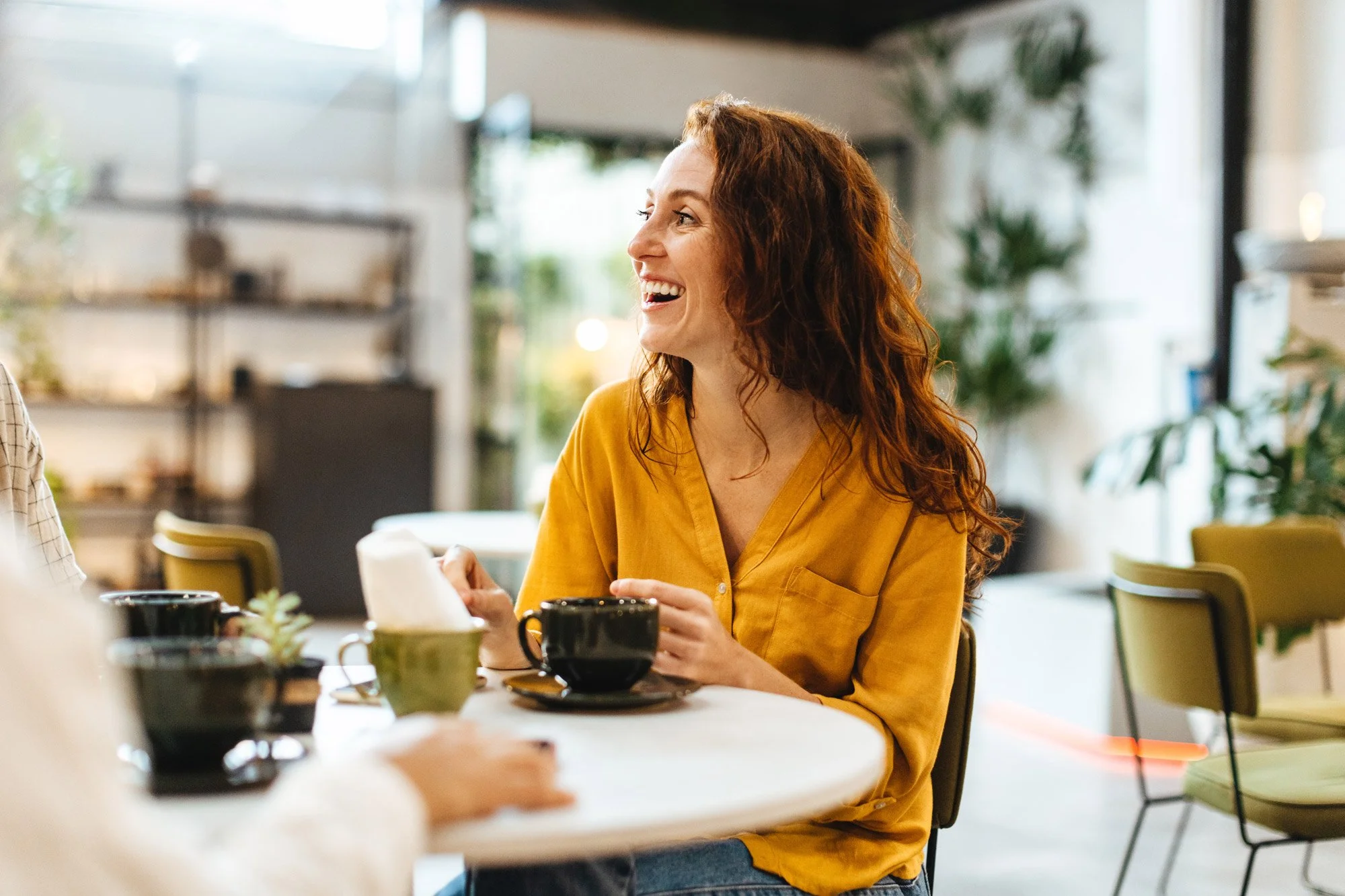A woman with curly red hair wearing a yellow blouse, smiling and talking at a cafe table with black cups and a tissue box.
