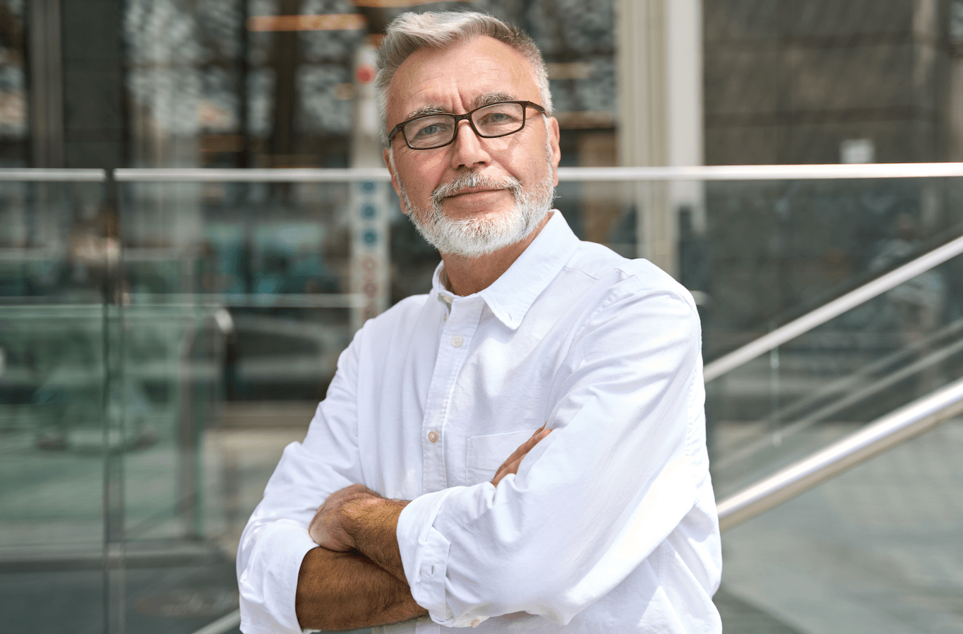 An older man with gray hair, beard, and glasses standing outdoors with arms crossed, wearing a white button-up shirt.