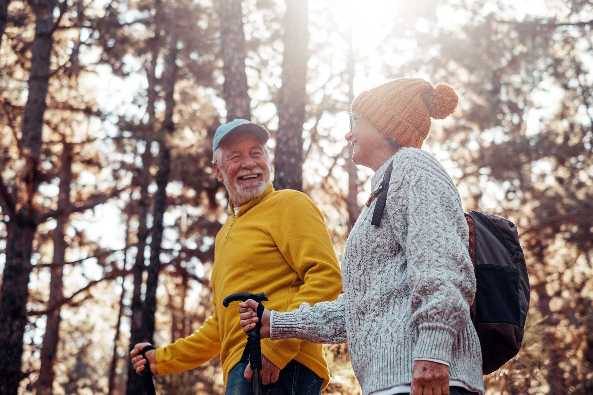 Two elderly people, a man wearing a yellow sweater and a woman wearing a gray knit sweater and a brown beanie, enjoy walking together in a sunlit forest.