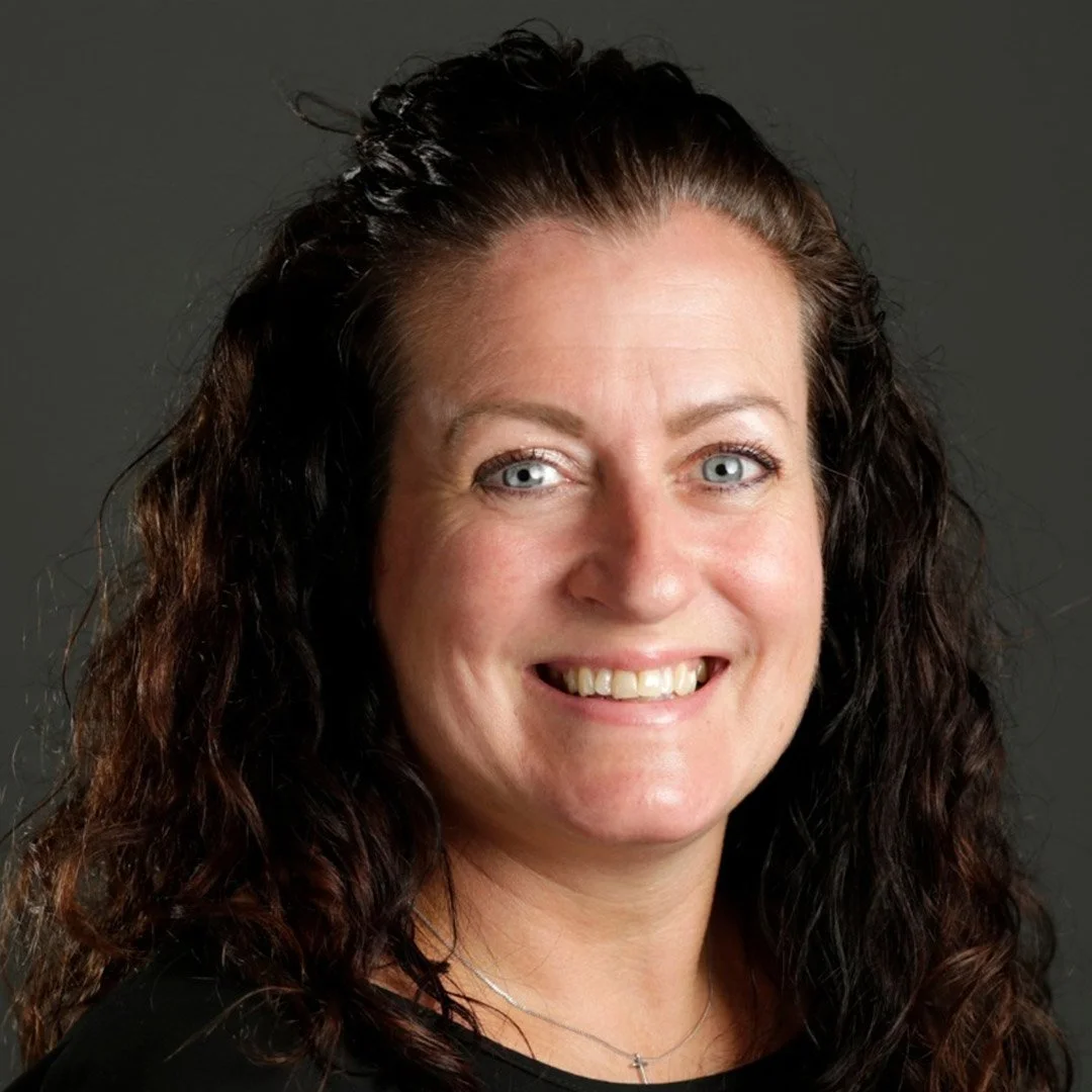 A woman with curly brown hair, light skin, and blue eyes smiling against a gray background.