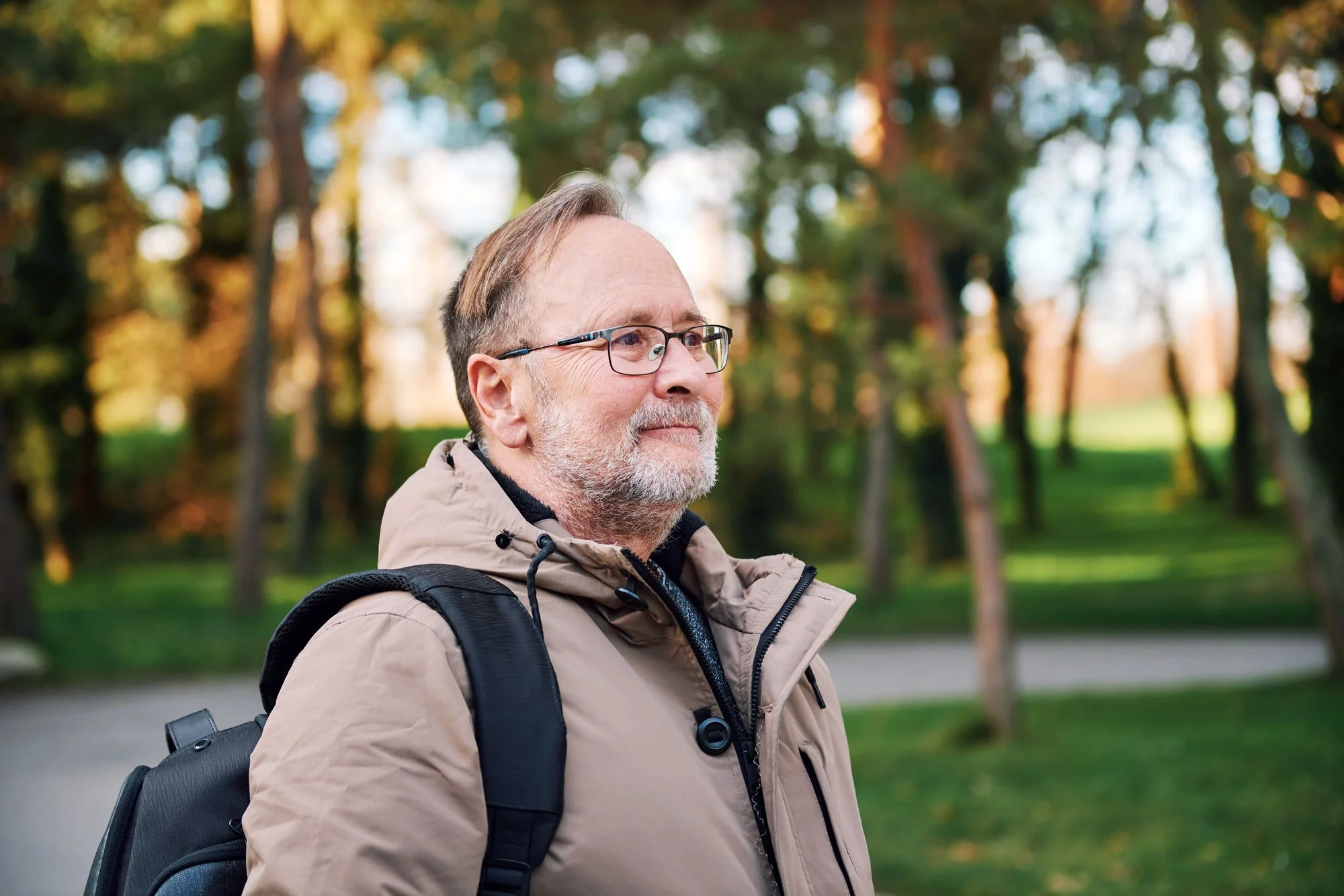 A mature man with glasses and a gray beard, wearing a beige jacket and carrying a backpack, stands outdoors in a park with green grass and trees, during what appears to be early morning or late afternoon.