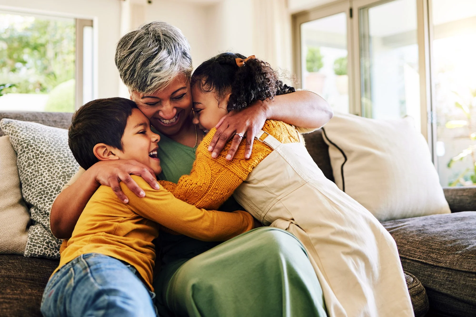 A happy multigenerational family hugging on a couch inside a bright living room with large windows and outdoor greenery in the background.