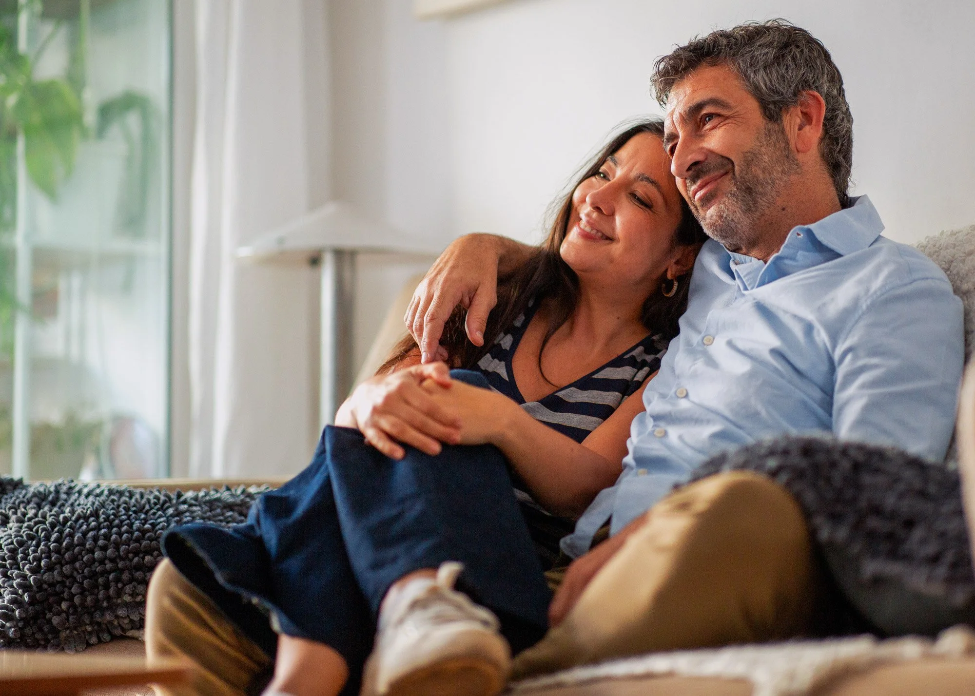 A middle-aged couple sitting on a sofa, smiling and embracing each other, in a cozy living room with natural light.