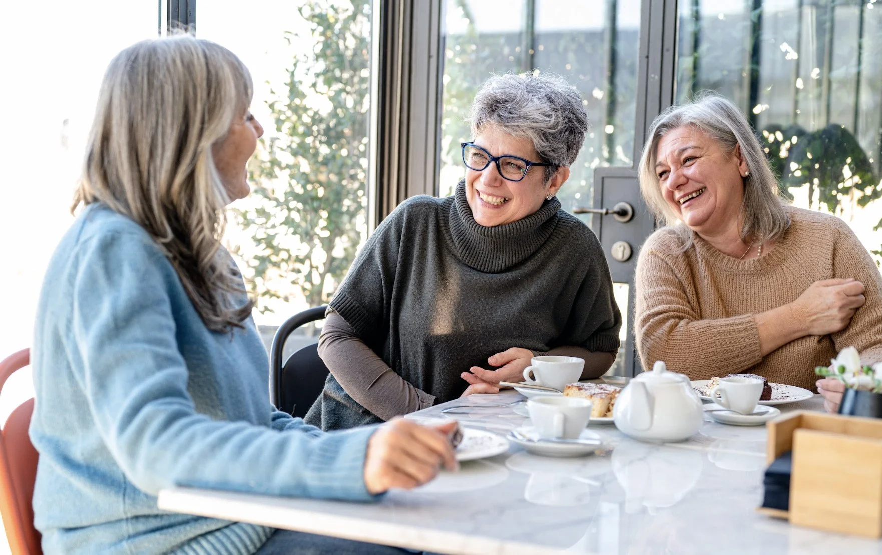 Three women smiling and laughing at a table with tea and cake during daytime.