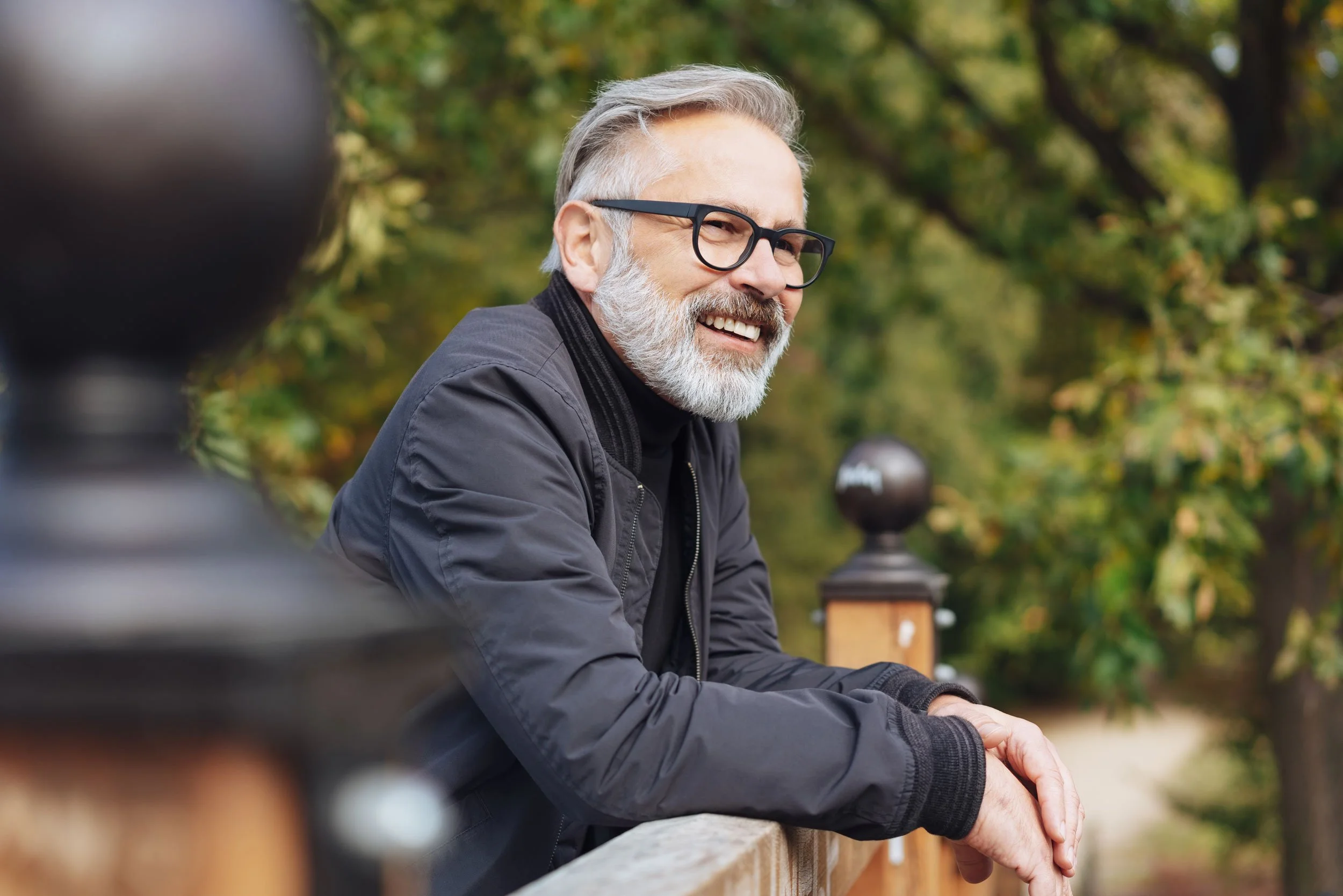 A smiling older man with gray hair, beard, and glasses sitting outdoors on a wooden railing, with greenery in the background.