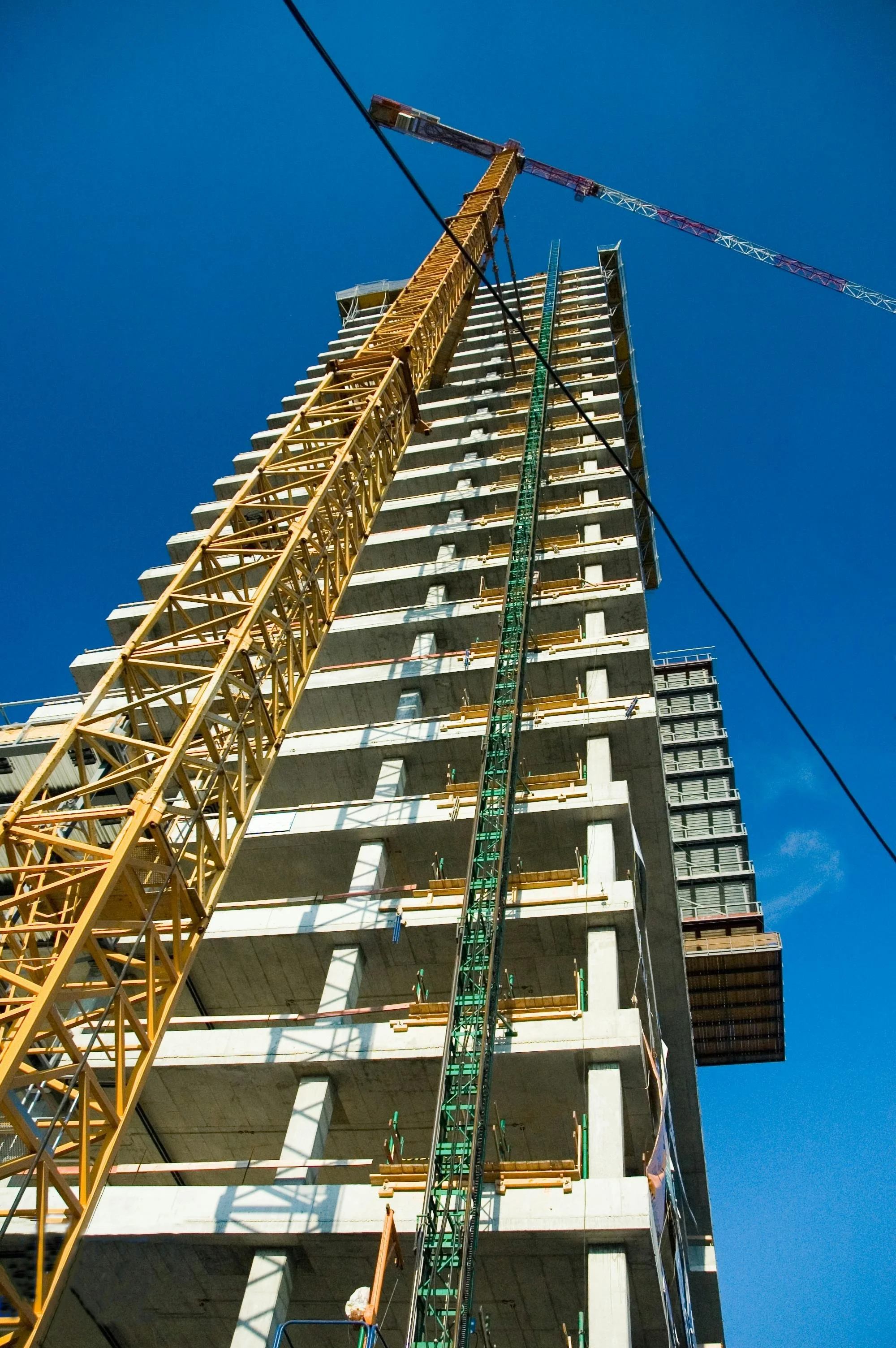 Construction site with a tall building under construction, a yellow tower crane, and a green construction elevator, against a clear blue sky.