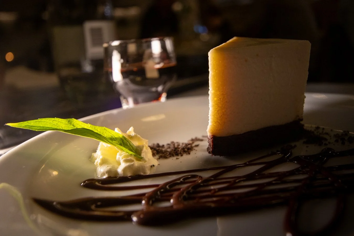 A slice of Bistro 129 Bailey's chocolate cheesecake on a white plate, garnished with a green herb and served with whipped cream and chocolate drizzle.