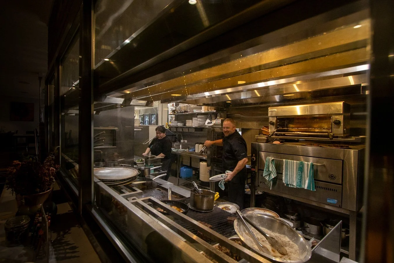 Two chefs are working in a commercial kitchen at Bistro 129 seen through an open window, with various pots, pans, and cooking equipment around them as they work.
