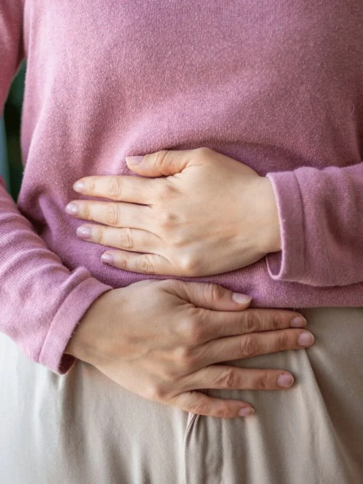 Deux mains posées sur le ventre d'une personne portant un pull rose et un pantalon beige.