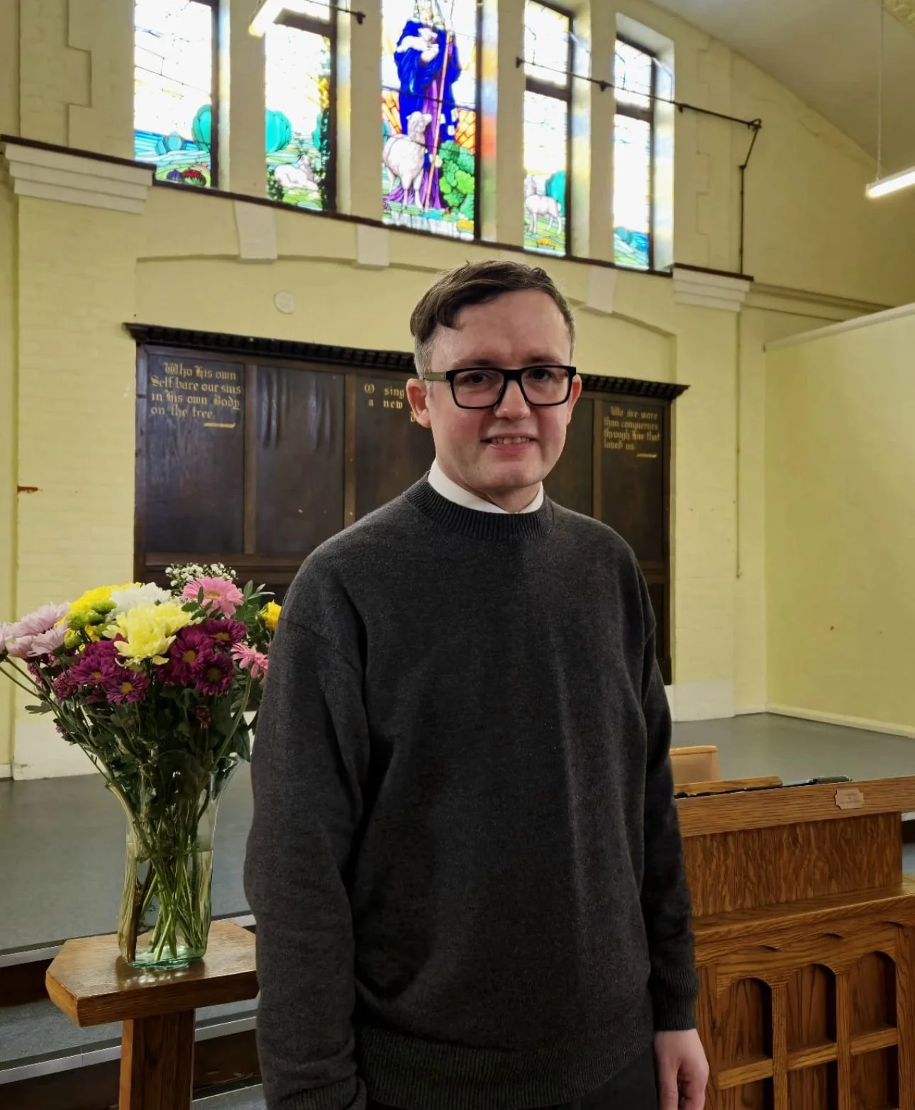 Young man wearing glasses and a dark sweater standing inside a church, with a church window mural, a wooden pulpit, and a vase of colorful flowers nearby.
