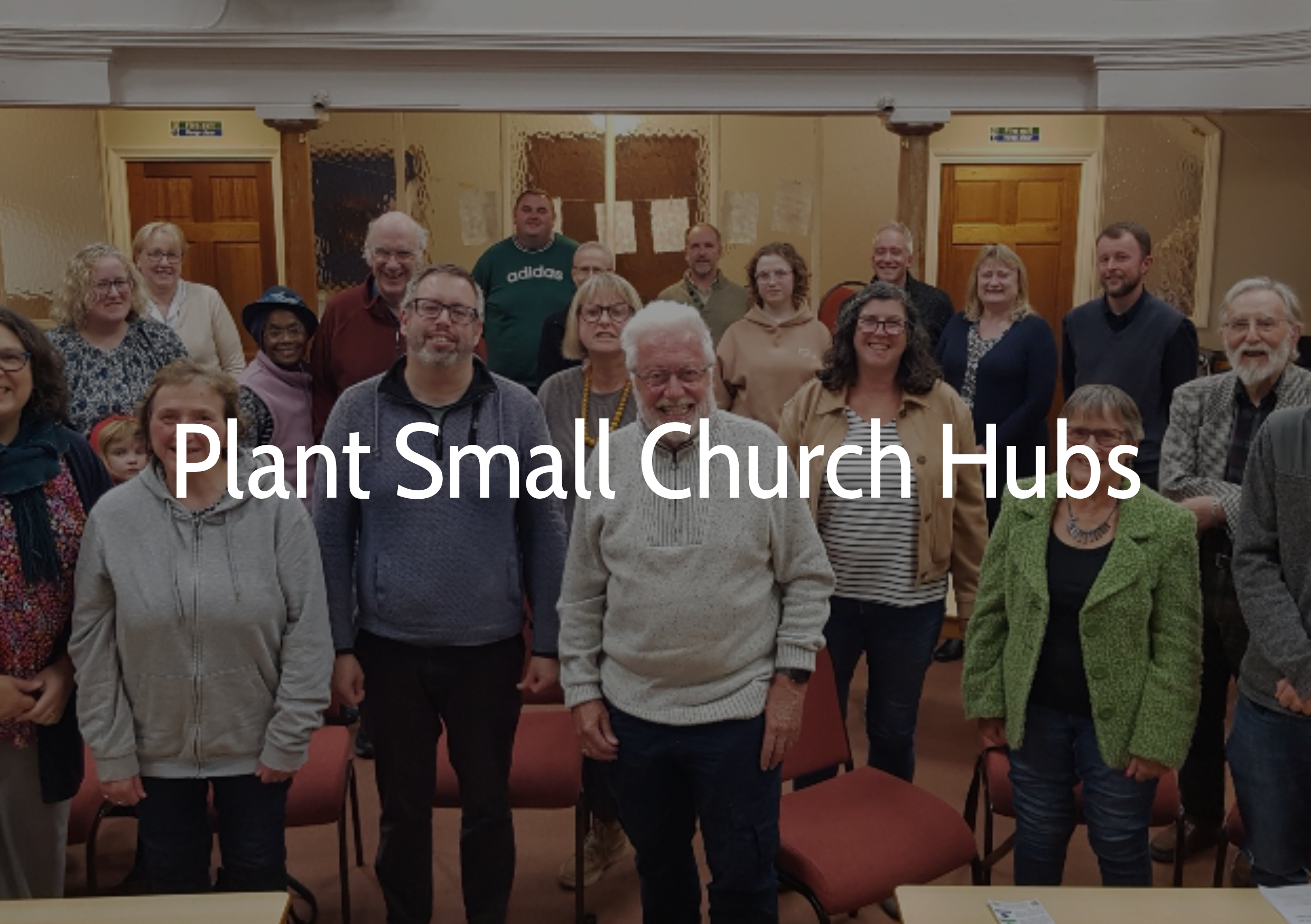 Group of diverse people smiling and standing together in a room with wooden doors and papers on the wall, labeled 'Plant Small Church Hubs'.