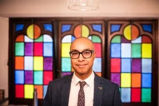 Man in a suit and glasses standing in front of stained glass windows.