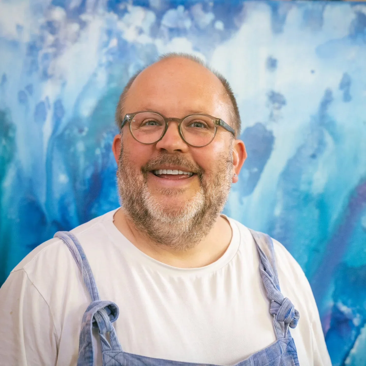 A smiling man with glasses, a beard, and a mustache, wearing a white t-shirt with blue suspenders, in front of a colorful abstract blue background.
