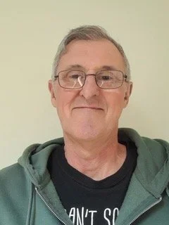 A middle-aged man with short gray hair, glasses, and a slight smile, wearing a dark t-shirt and a green hoodie, standing against a light-colored wall.