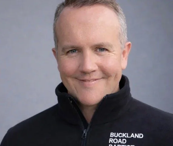Portrait of a smiling middle-aged man wearing a black jacket with a zip and the words 'BUCKLAND ROAD