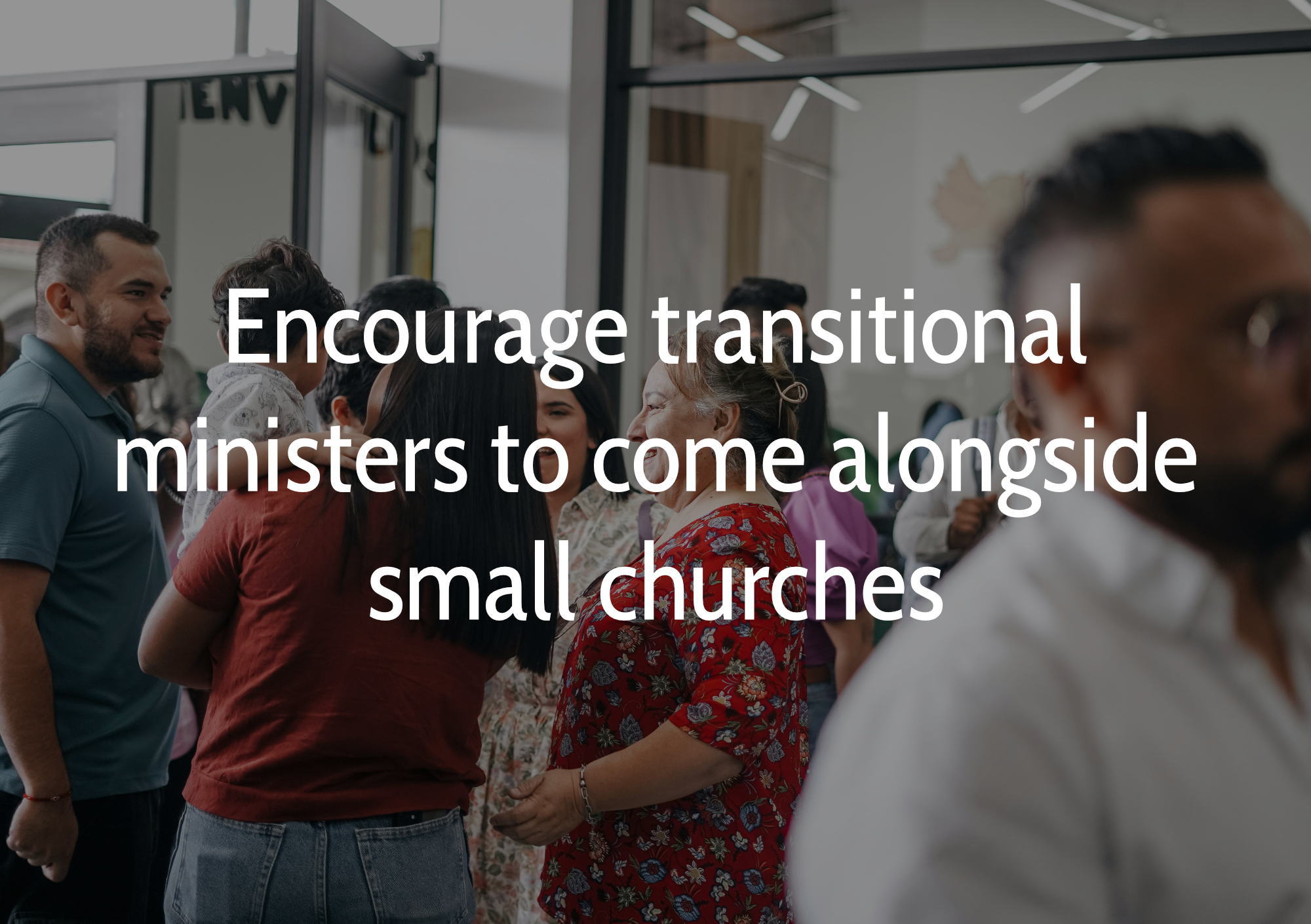 A diverse group of people, including men and women, gathered inside a modern building with glass walls, engaged in conversations and handshakes, suggesting a community or church gathering.