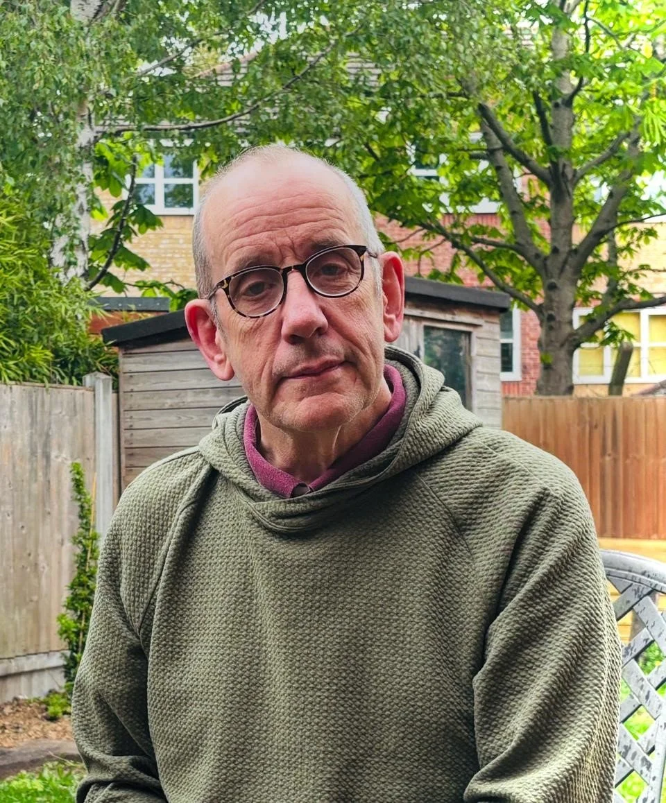An older man wearing glasses and a green textured hoodie standing outdoors in a garden with trees, a wooden fence, and a shed in the background.