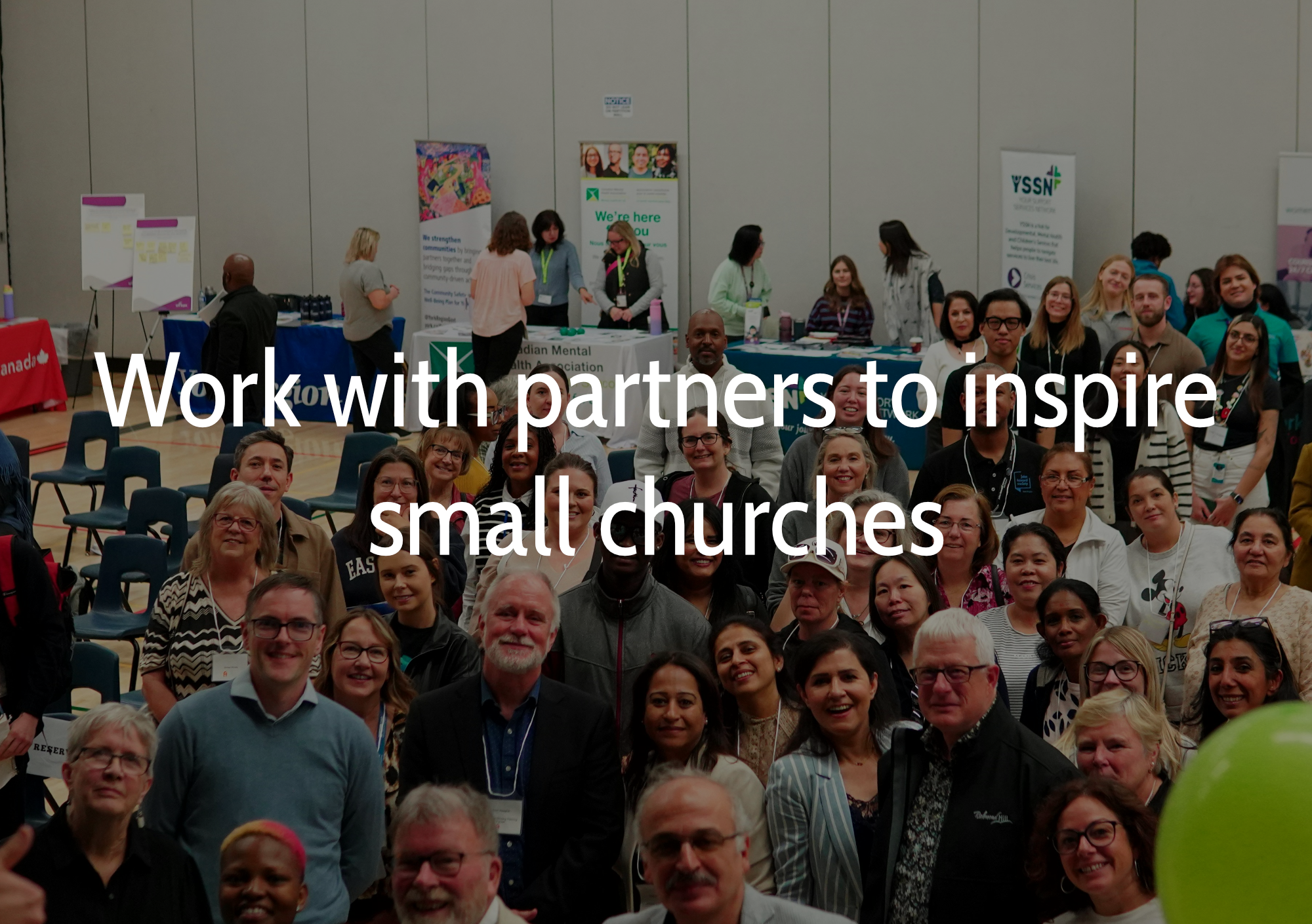 A group of diverse people gathered at a community event or conference, some smiling for the camera with booths and banners in the background. Text reads 'work with partners to inspire small churches'.