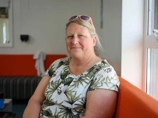 A middle-aged woman with blonde hair wearing sunglasses and a floral shirt sitting on an orange bench near a window.
