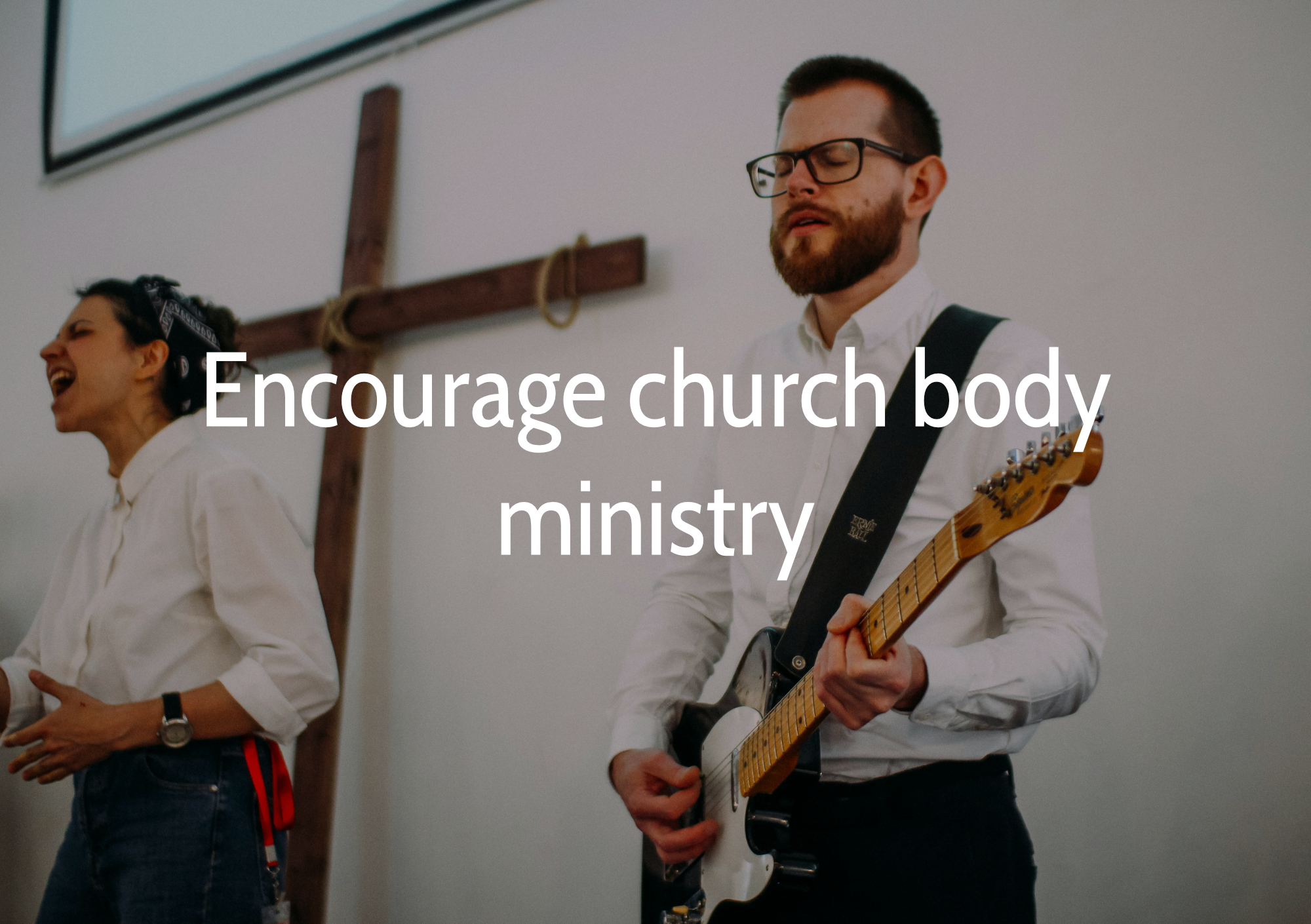A man with glasses and a beard playing an electric guitar during a church service, with a woman singing and a cross in the background.