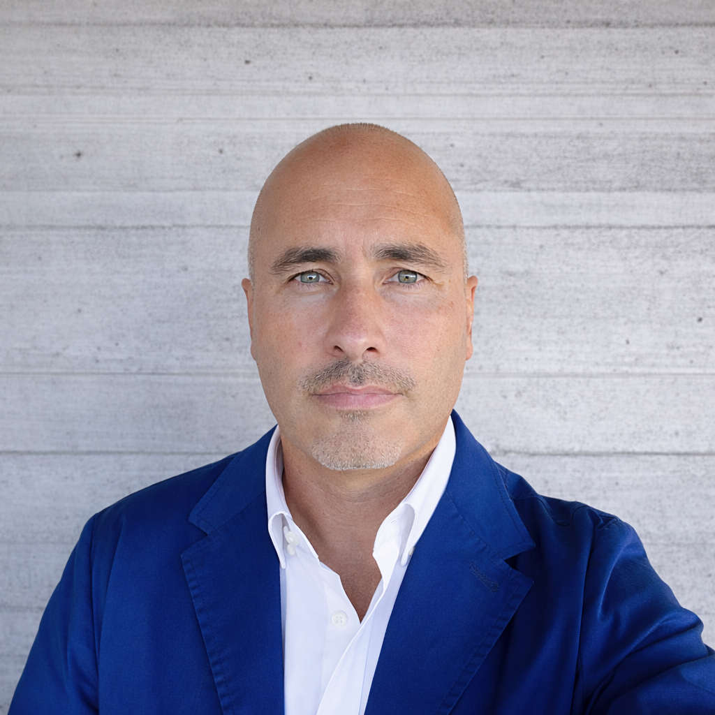 A close-up portrait of a bald man with light facial hair, wearing a white shirt and a dark blue blazer, standing in front of a gray wooden wall.